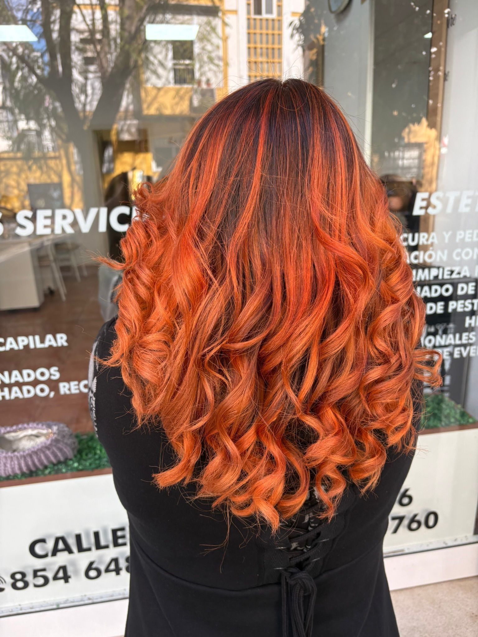Vista posterior de una larga cabellera rizada de color naranja rojizo reflejada en el escaparate de una peluquería.