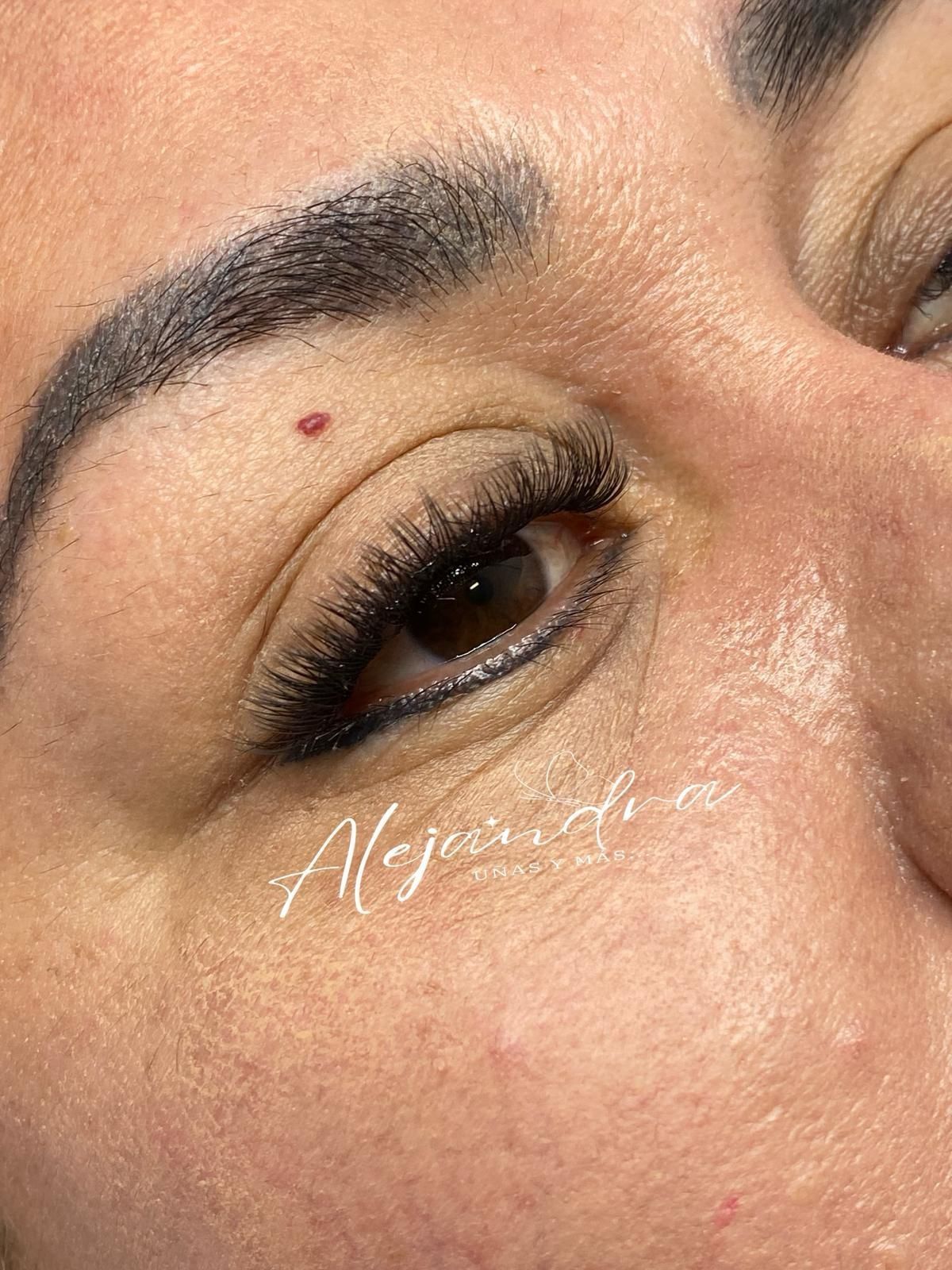 Primer plano del ojo de una persona con delineador de ojos, pestañas largas y un pequeño lunar en la zona de la ceja.