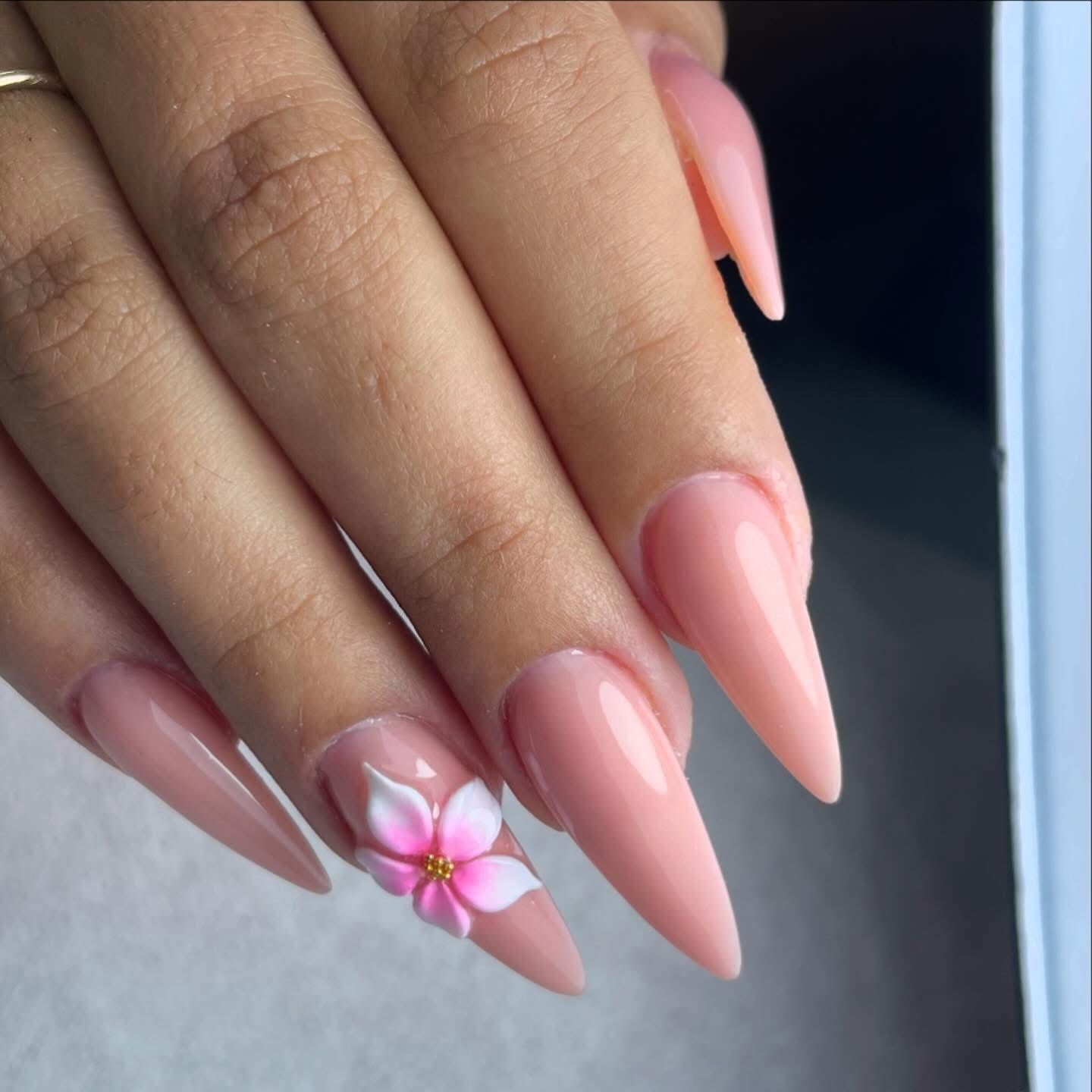 Mano con uñas largas de color rosa almendrado y una pequeña flor blanca como adorno en las uñas.