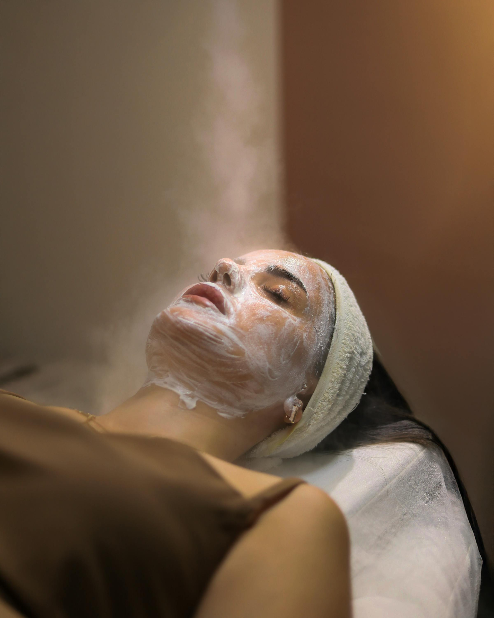 Mujer recibiendo un tratamiento facial con una mascarilla blanca en el rostro, recostada en una camilla de spa.