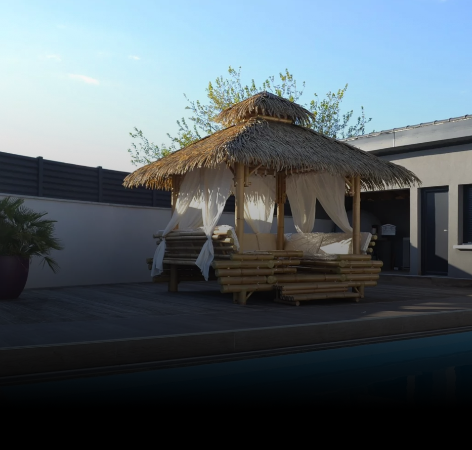 Belvédère en bambou avec toit de chaume, rideaux blancs, sur patio près du bâtiment, ciel bleu.