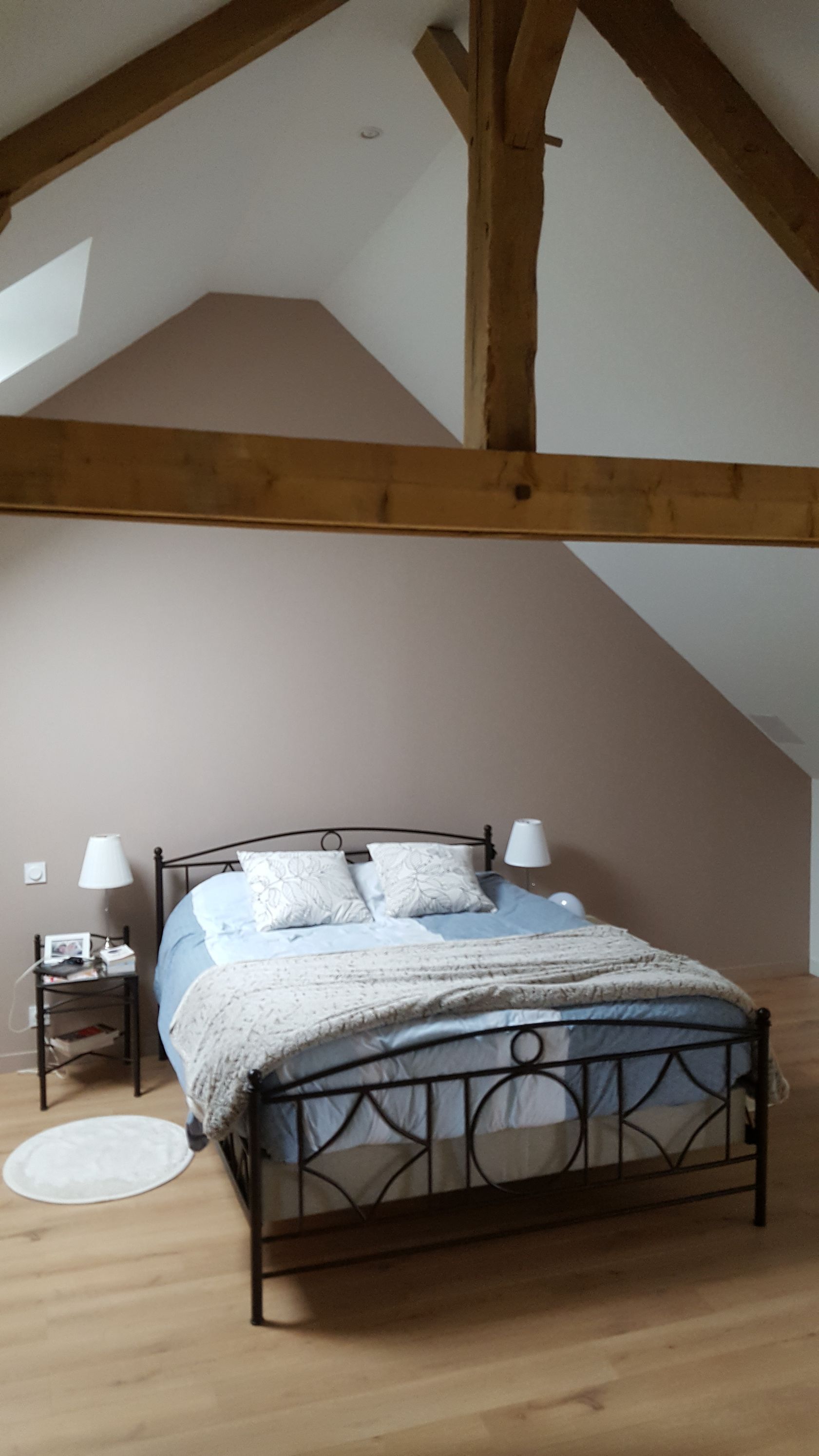 Chambre avec un lit en métal, un mur rose pâle, des poutres en bois et un parquet.