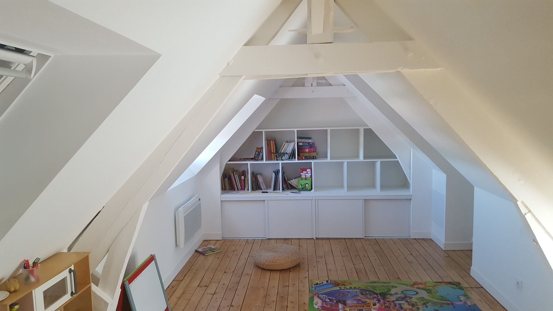 Chambre mansardée avec poutres blanches, étagères intégrées, parquet et tapis coloré.