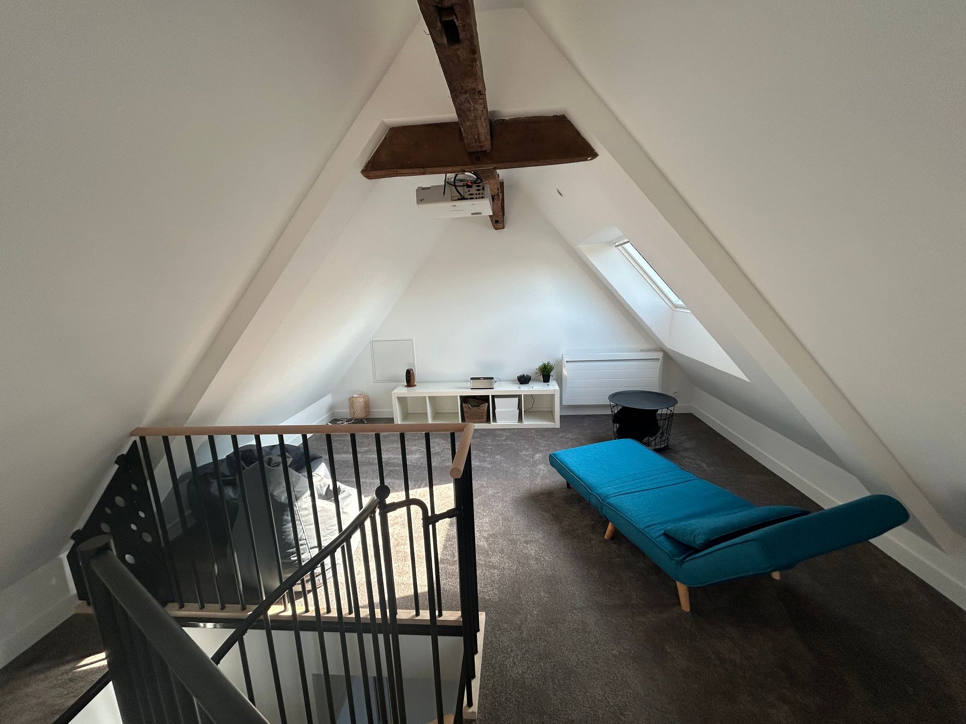 Chambre mansardée avec futon, escalier et poutres apparentes. Murs blancs et plafond en pente.