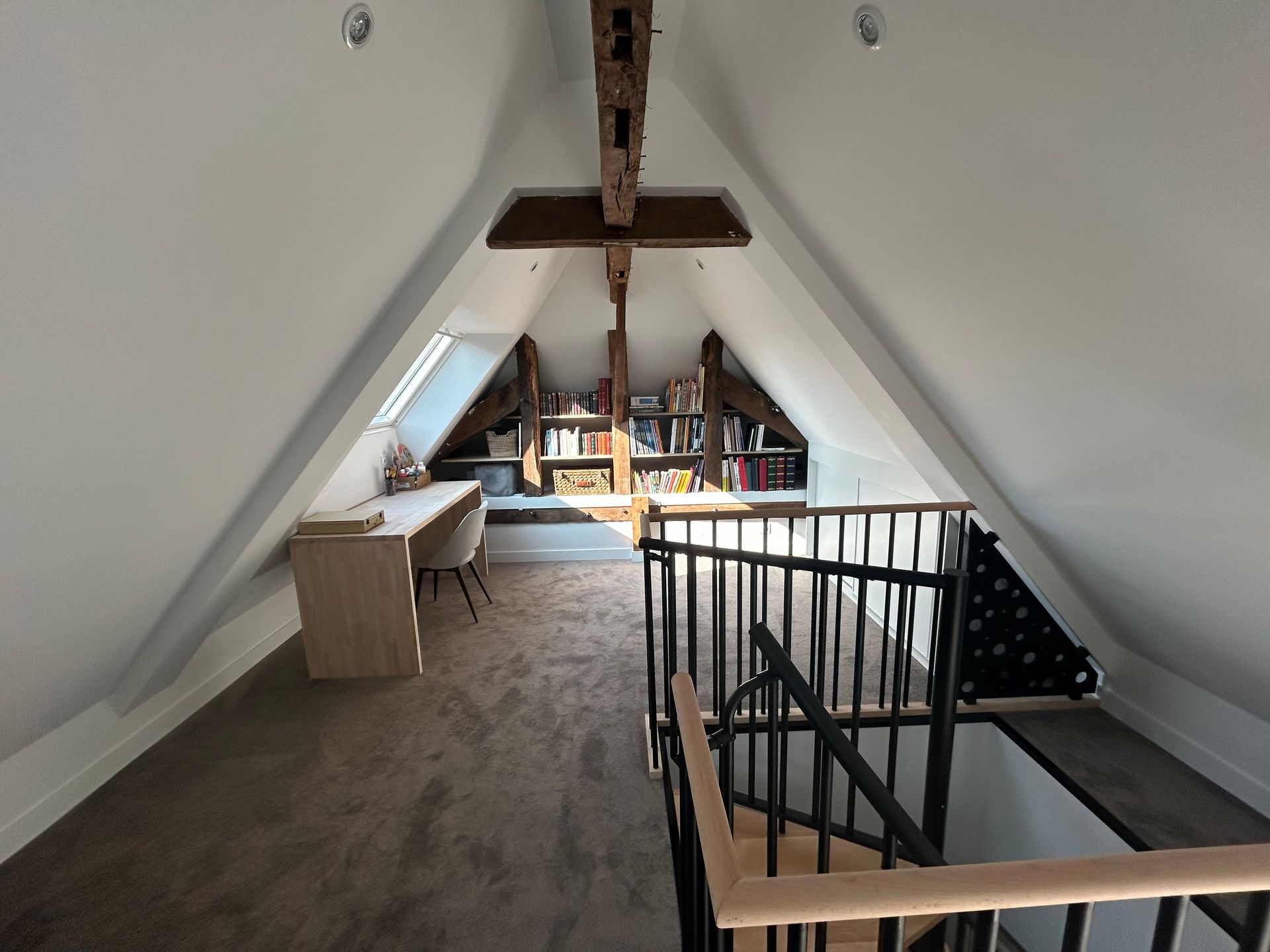 Bibliothèque mansardée avec murs en pente, bureau, bibliothèque et escalier avec rampe noire.