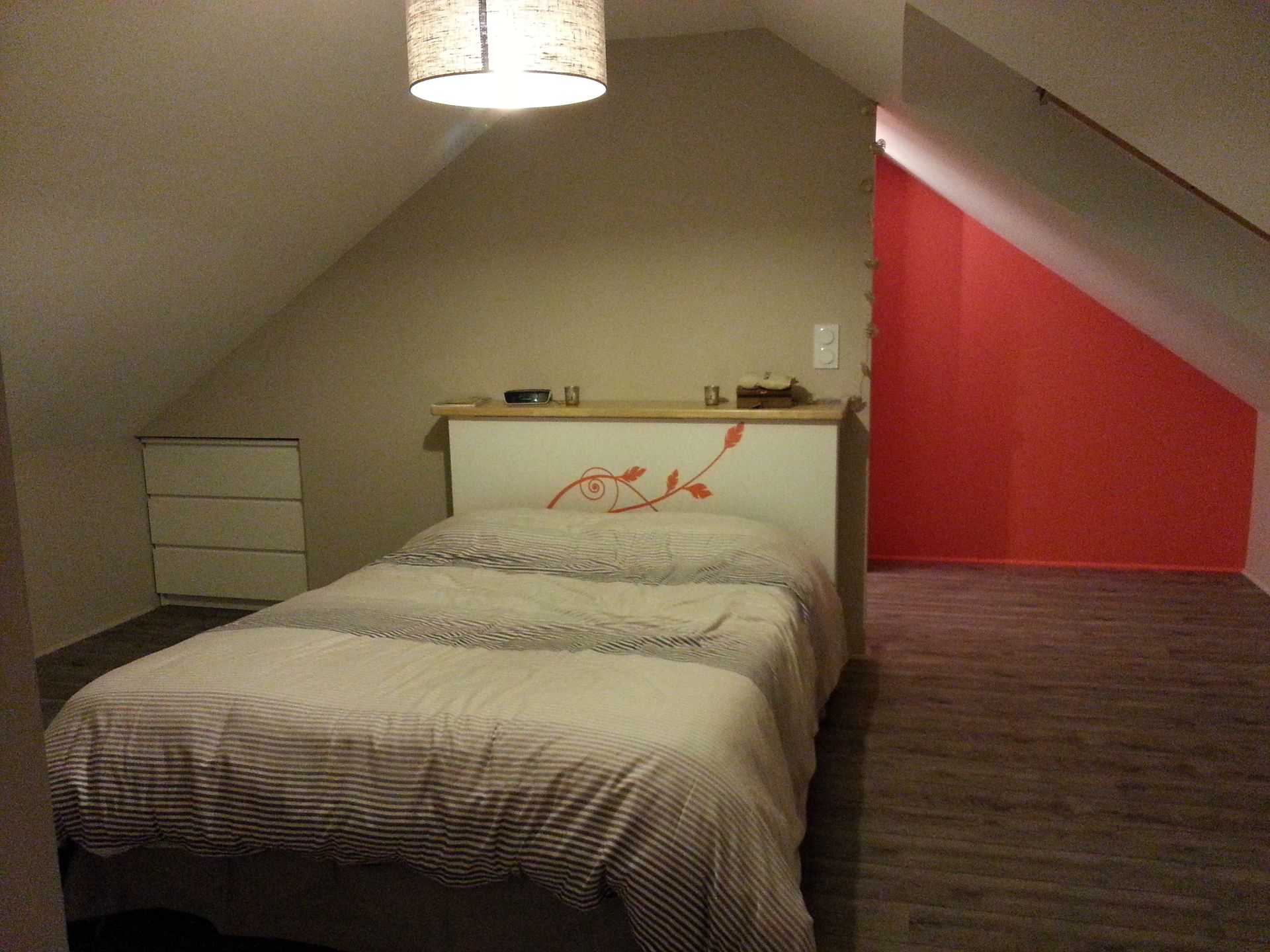 Chambre mansardée avec murs en pente, un lit, une commode et un mur d'accent rouge.