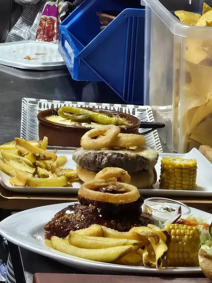 Platos de hamburguesas, papas fritas, aros de cebolla y maíz sobre una mesa de restaurante con condimentos y una mampara azul.