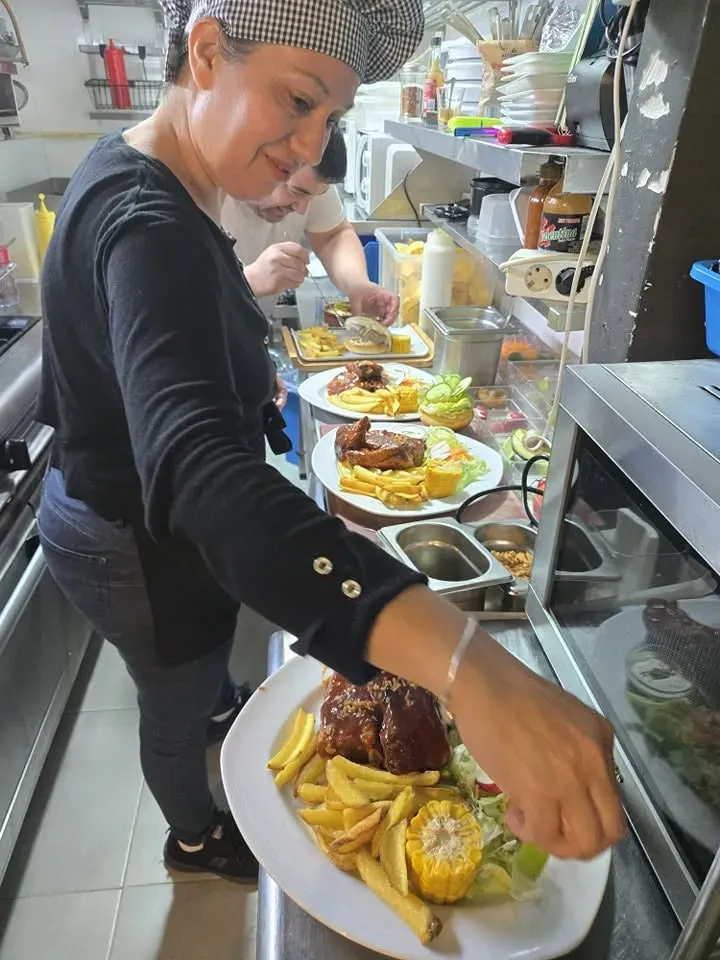 Una persona coloca platos de comida en un mostrador concurrido, con pollo frito y plátanos fritos.
