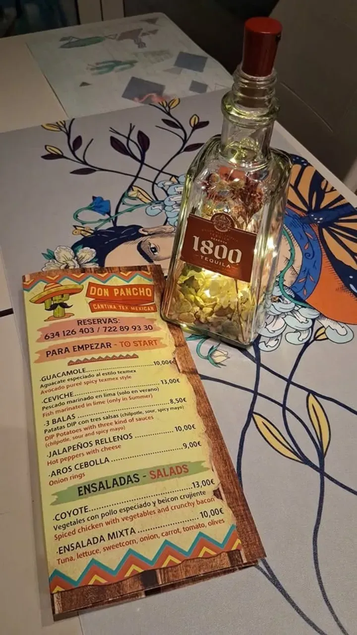 Una botella de tequila 1800 junto a una colorida carta de bebidas sobre un mantel estampado.