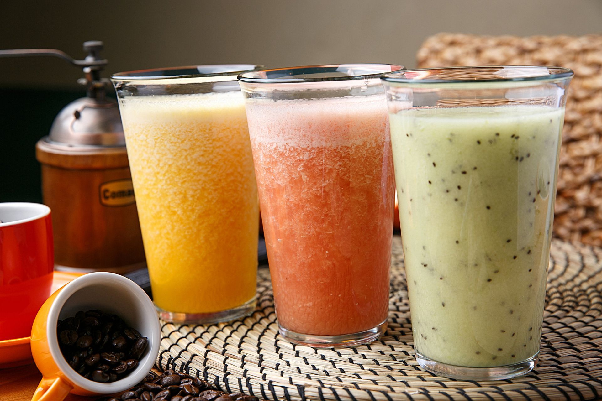 Trois smoothies aux fruits dans des verres transparents – jaune, rouge et vert – sur une table avec des grains de café et un moulin.
