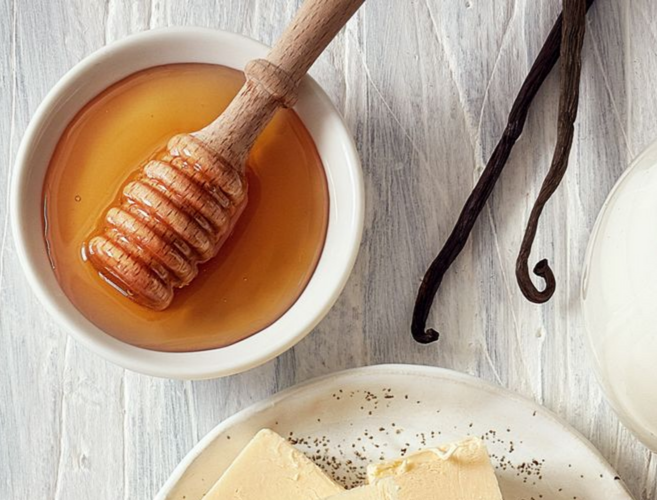 Du miel dans un bol avec une louche, des gousses de vanille, du beurre et un pot à lait sur une surface en bois blanc.