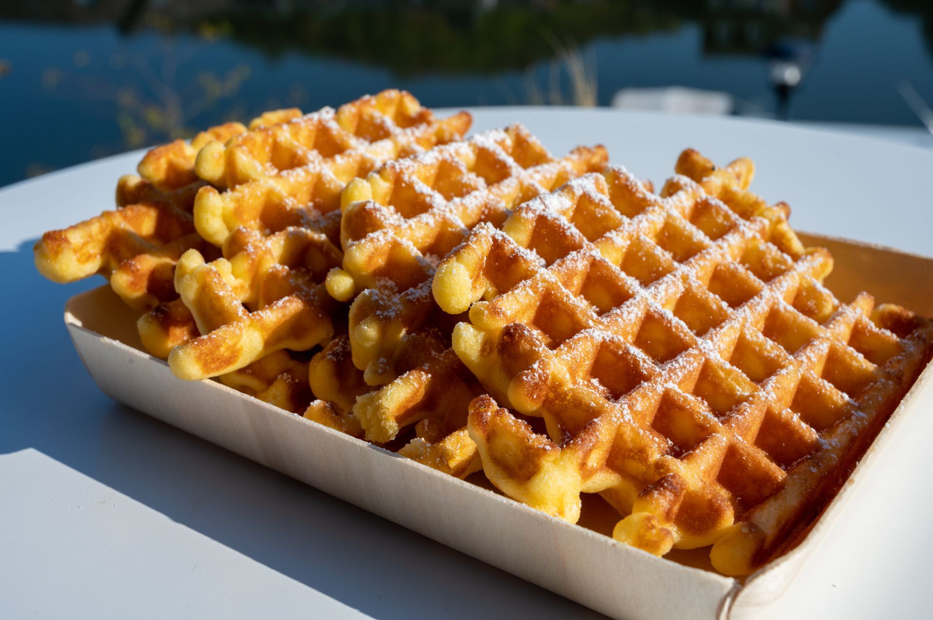 Pile de gaufres dorées sur un plateau en bois, saupoudrées de sucre glace, à l'extérieur au soleil.