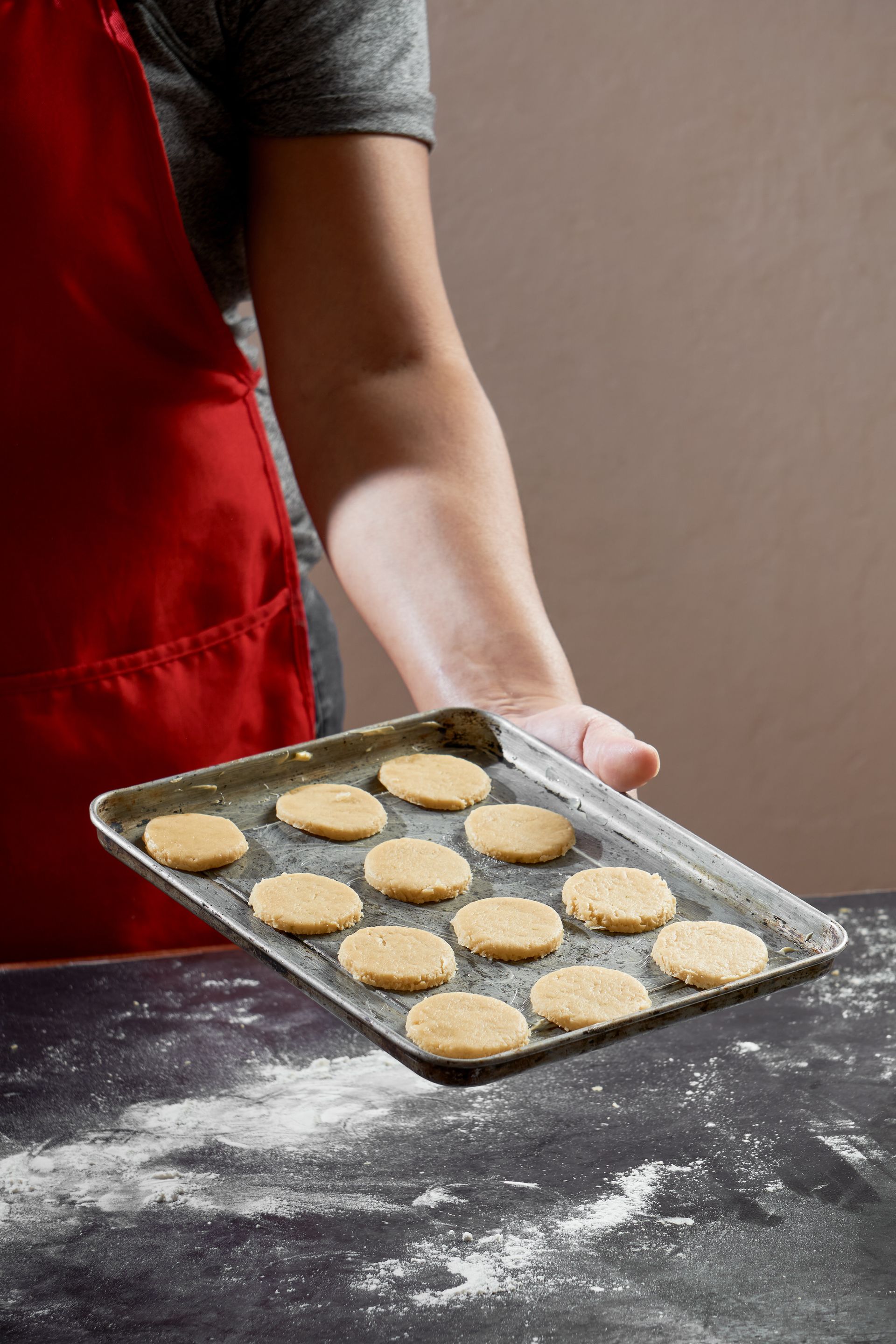 Personne tenant une plaque à pâtisserie de biscuits ronds et dorés, avec de la farine saupoudrée sur un plan de travail.