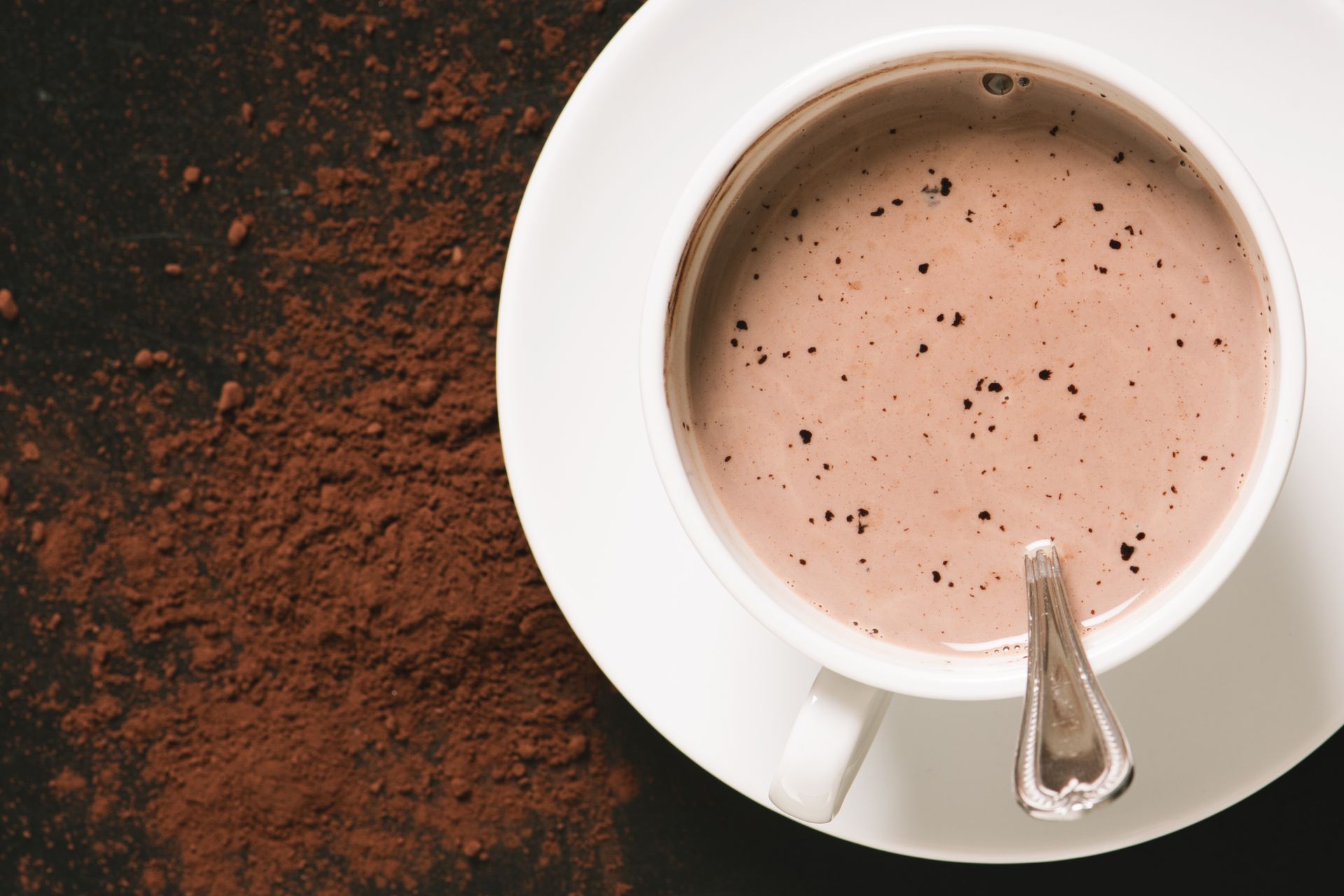 Chocolat chaud dans une tasse blanche avec une cuillère, sur une soucoupe blanche ; de la poudre de cacao se répand à proximité sur une surface sombre.