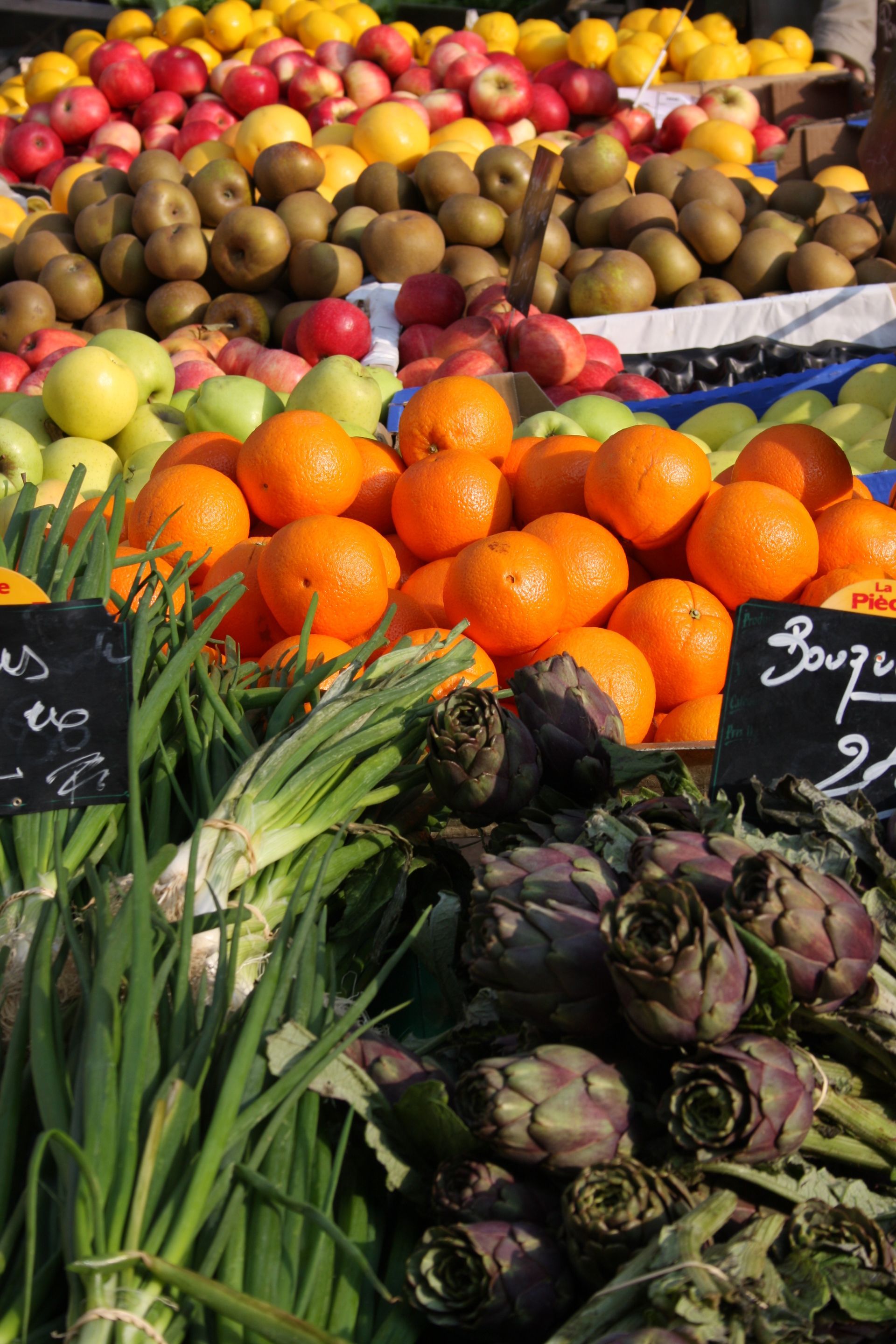 Produits frais exposés sur un étal de marché : oranges, artichauts, oignons verts, pommes, citrons et kiwis.