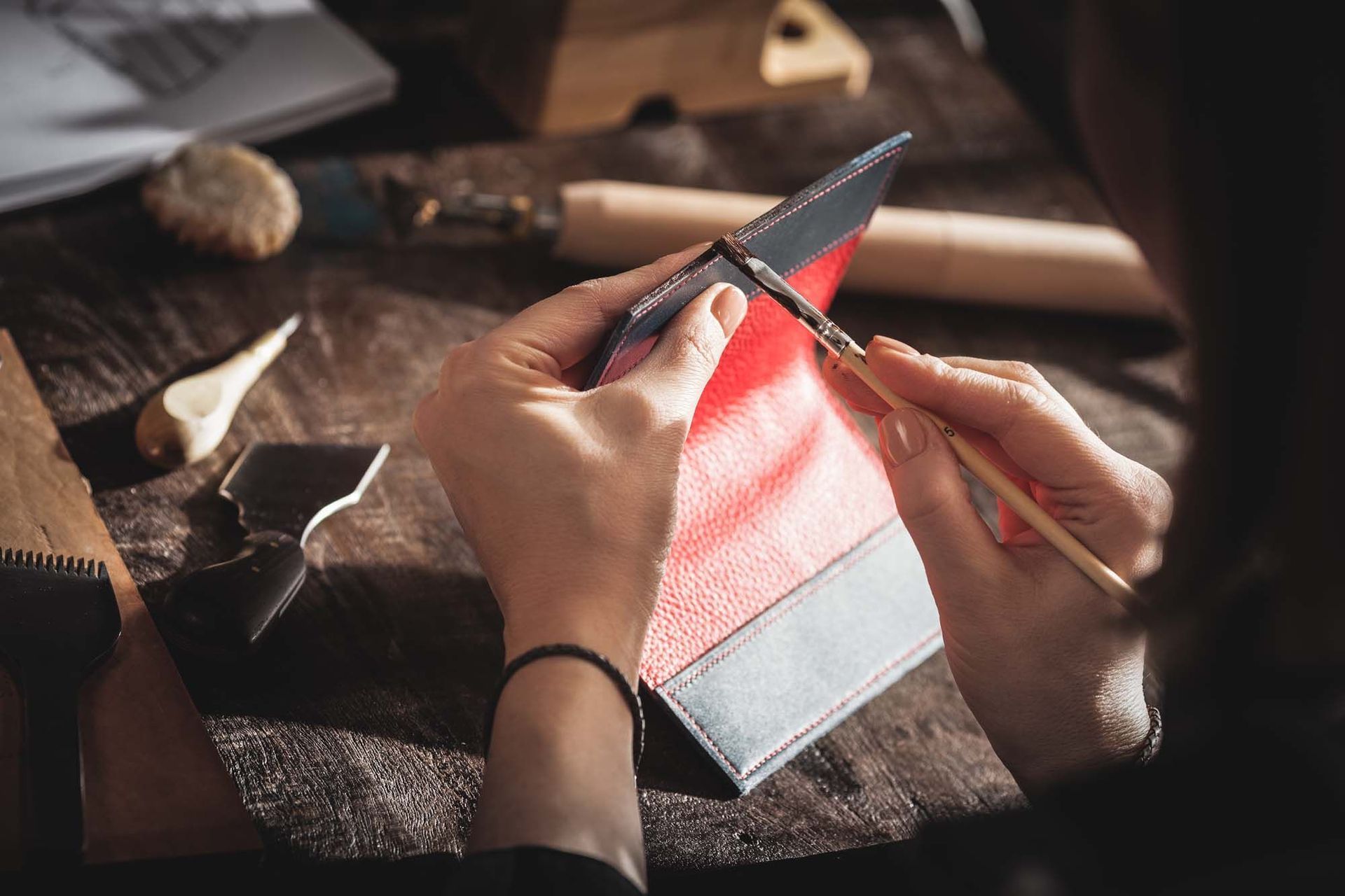 Hände bemalen den Rand eines Lederartikels auf einer hölzernen Arbeitsfläche; im Hintergrund sind Werkzeuge zu sehen.