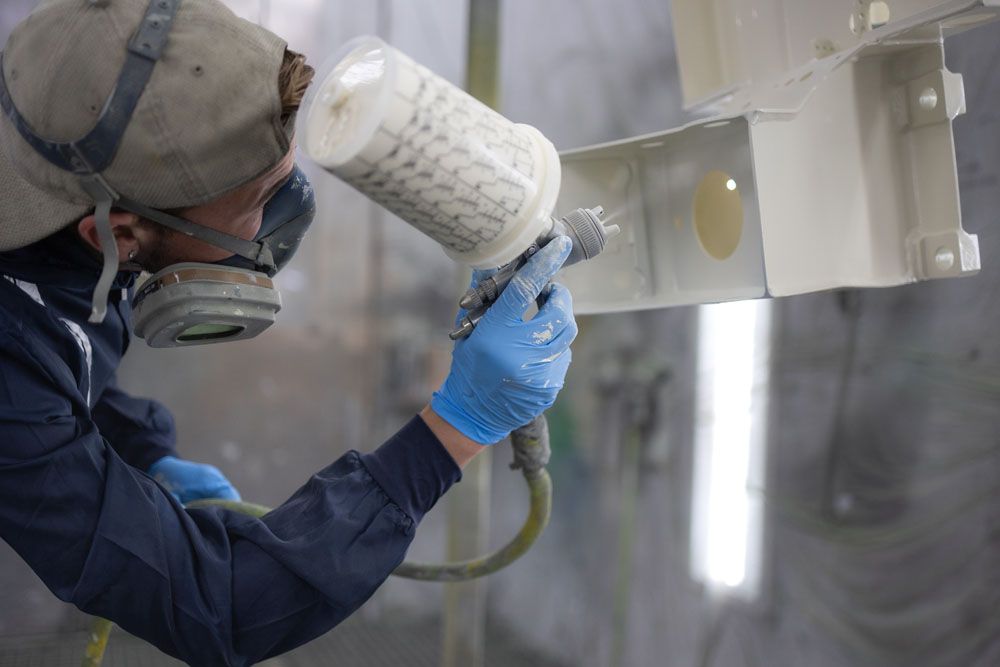 Ein Mann mit Maske und Handschuhen bemalt ein Stück Metall mit Sprühfarbe.
