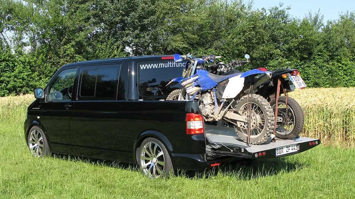 Ein schwarzer Lieferwagen mit zwei Dirtbikes auf der Ladefläche ist auf einer Wiese geparkt.