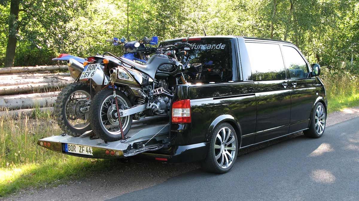 Ein schwarzer Lieferwagen mit zwei Motorrädern auf der Ladefläche steht am Straßenrand.