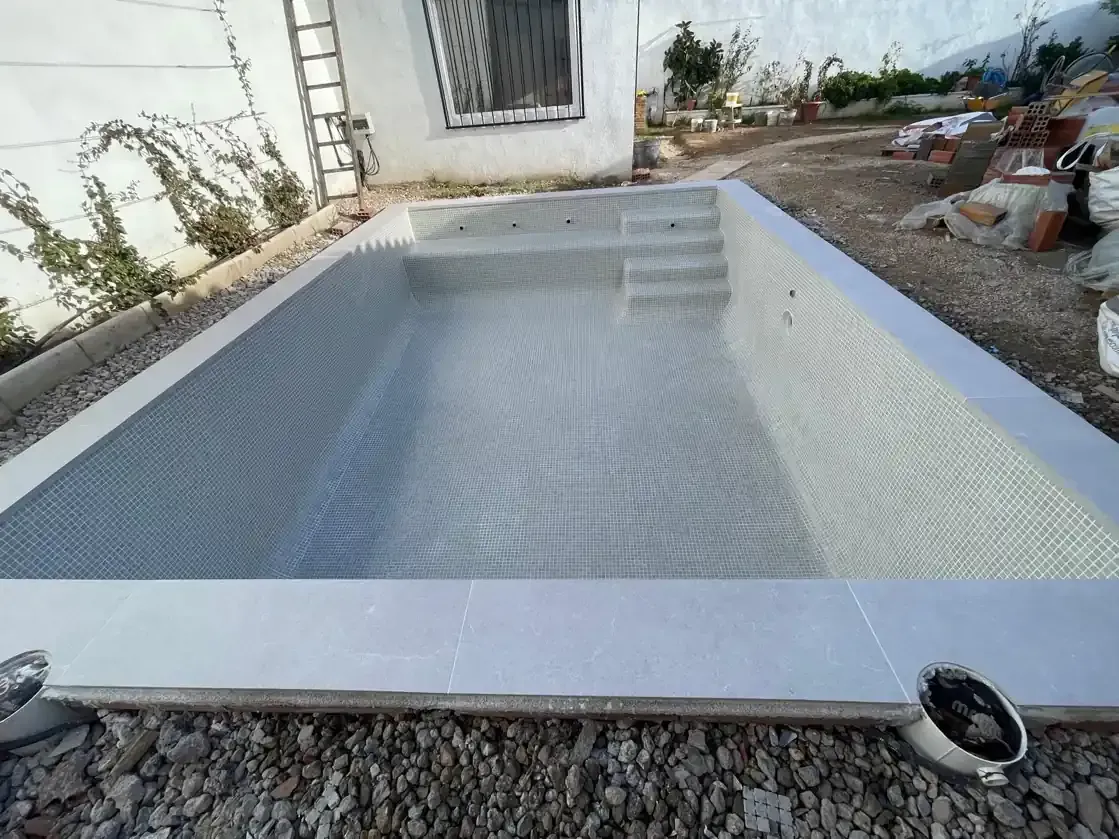 Piscina rectangular vacía en construcción junto a una casa blanca, rodeada de grava.