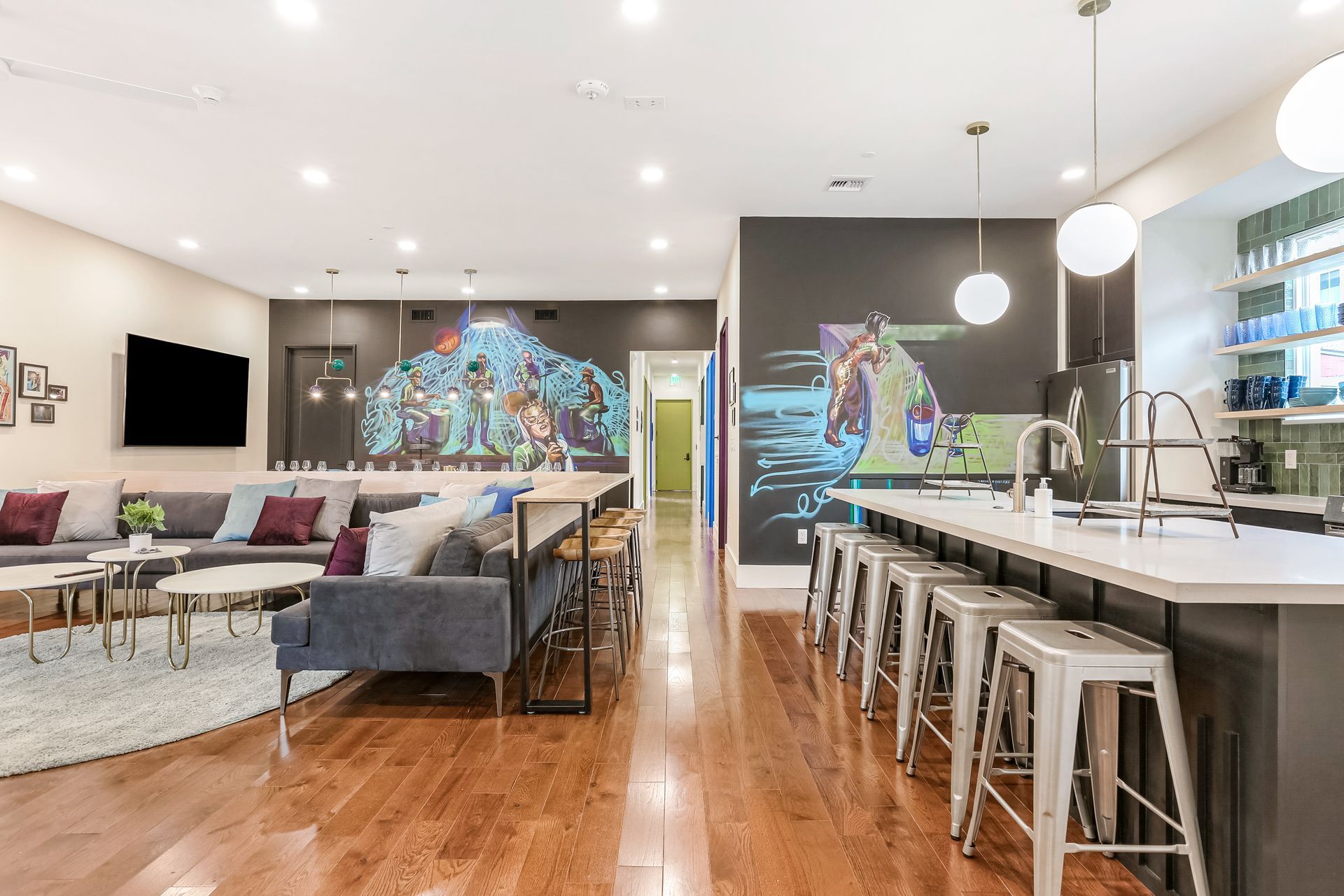 A living room with a couch , stools , a kitchen and a large painting on the wall.