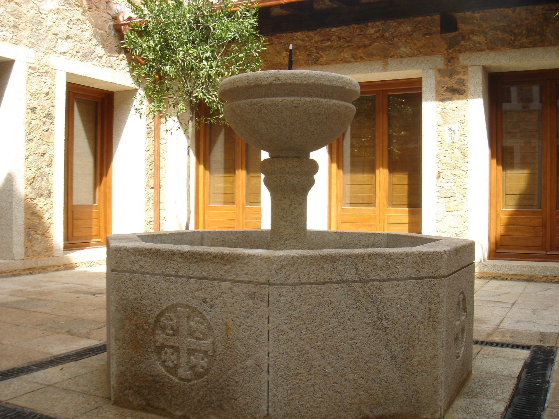 En un patio con muros de piedra clara y puertas de madera, se encuentra una fuente de piedra con base octogonal y parte superior escalonada.