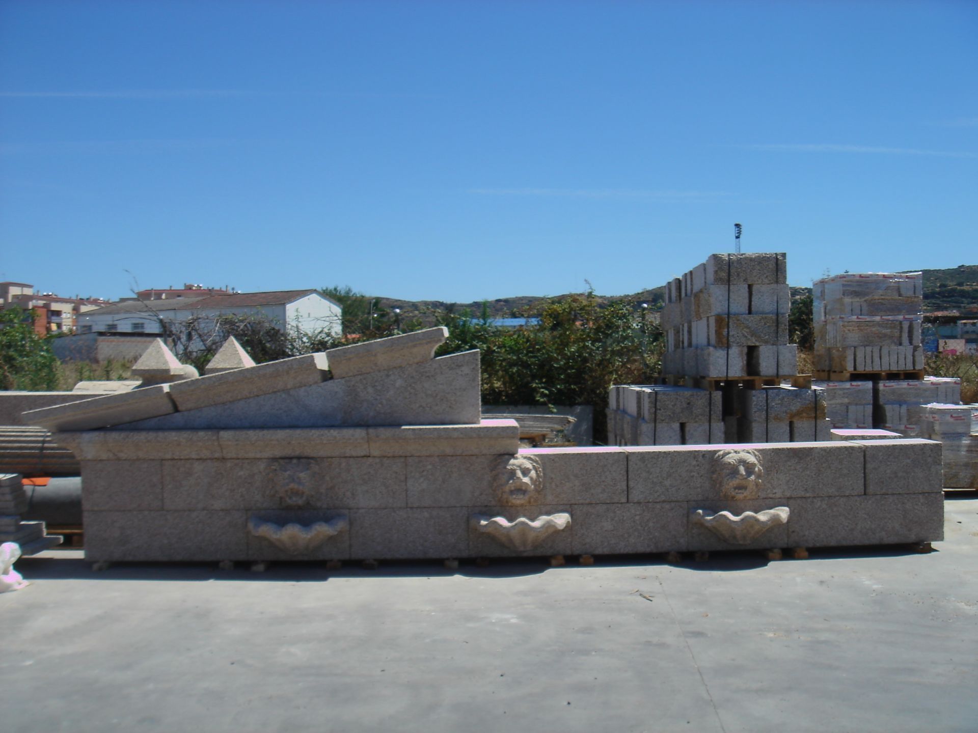 Una fuente de piedra con tres caños tallados en forma de cabeza de león se alza en un patio de piedra al aire libre, bajo un cielo azul despejado.