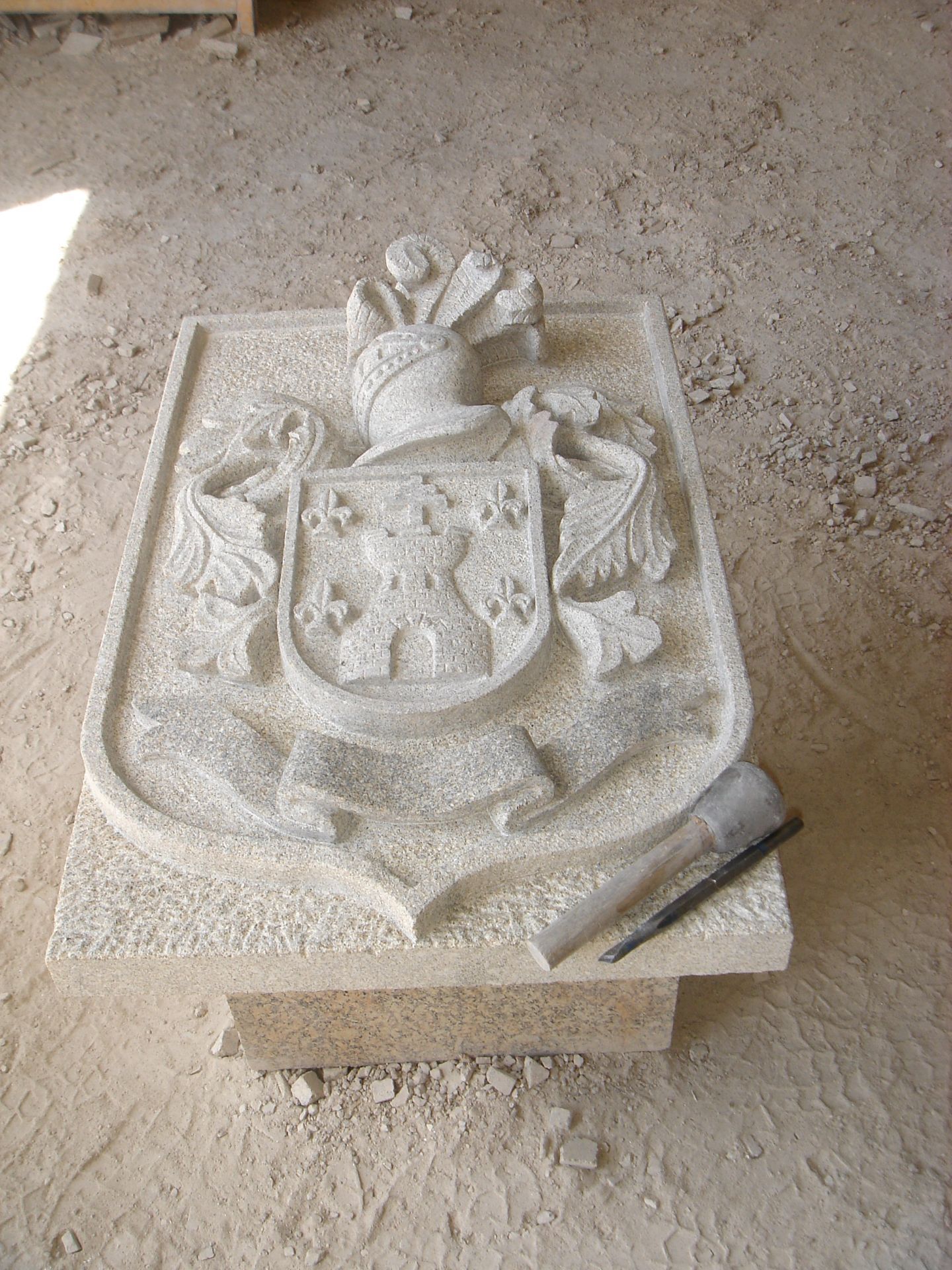 Una escultura de piedra que representa un escudo familiar con un castillo central y flores de lis, colocada sobre un pedestal con herramientas de cantero.