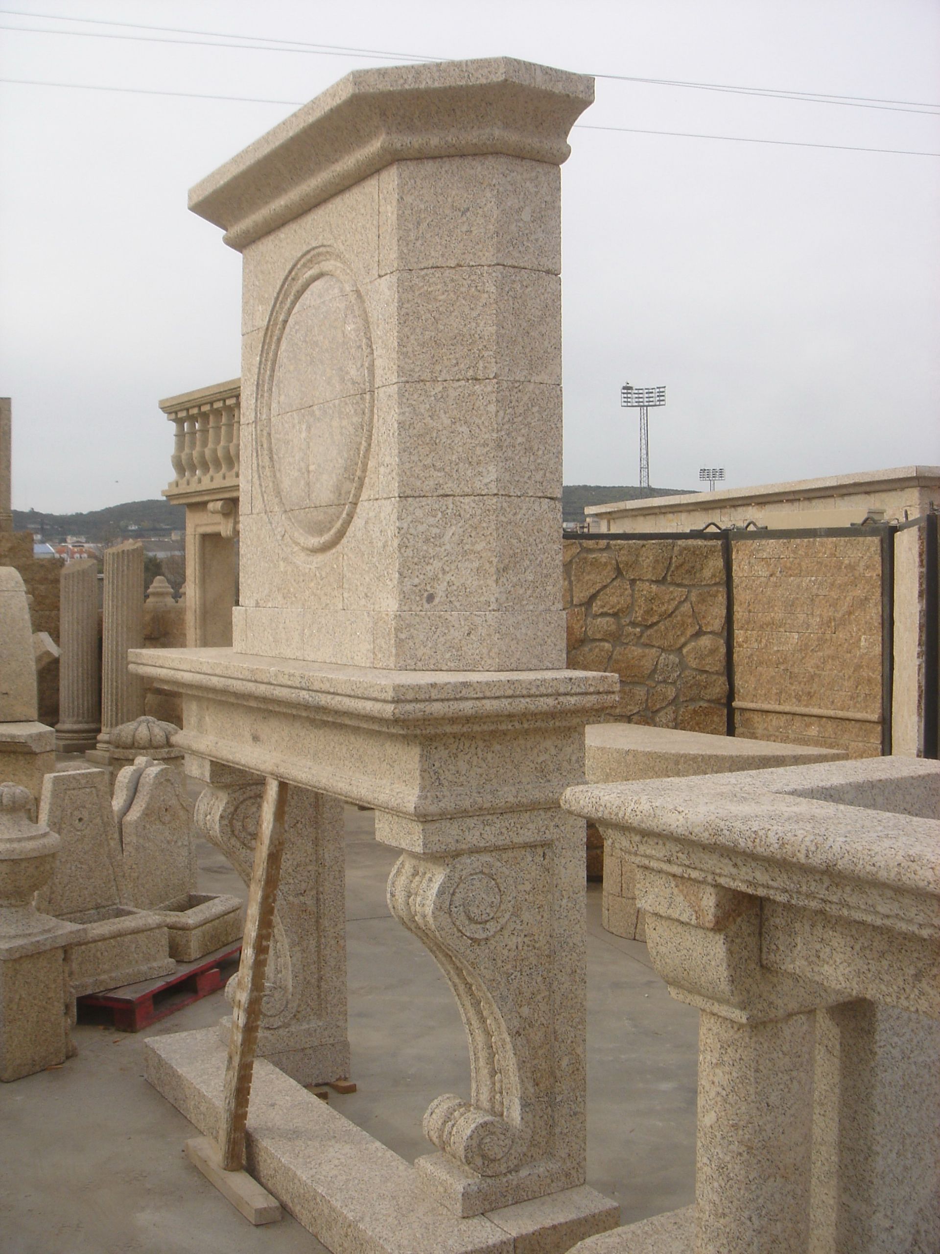 Una repisa de chimenea alta, de piedra clara y con una elaborada talla circular, ubicada en un taller de piedra al aire libre.