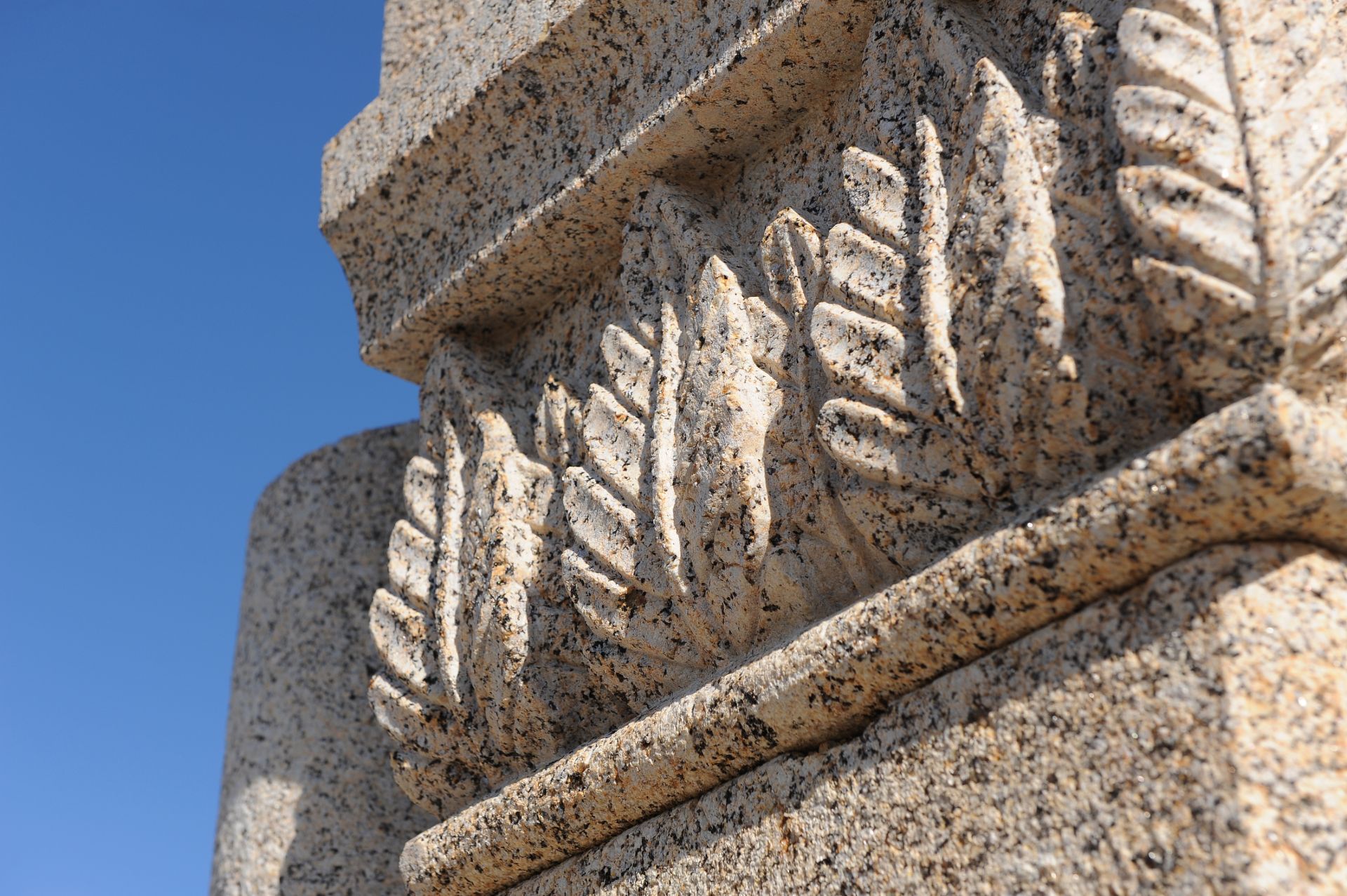 Escultura arquitectónica de granito con forma de hojas de helecho sobre un pilar de piedra, con un cielo azul despejado de fondo.