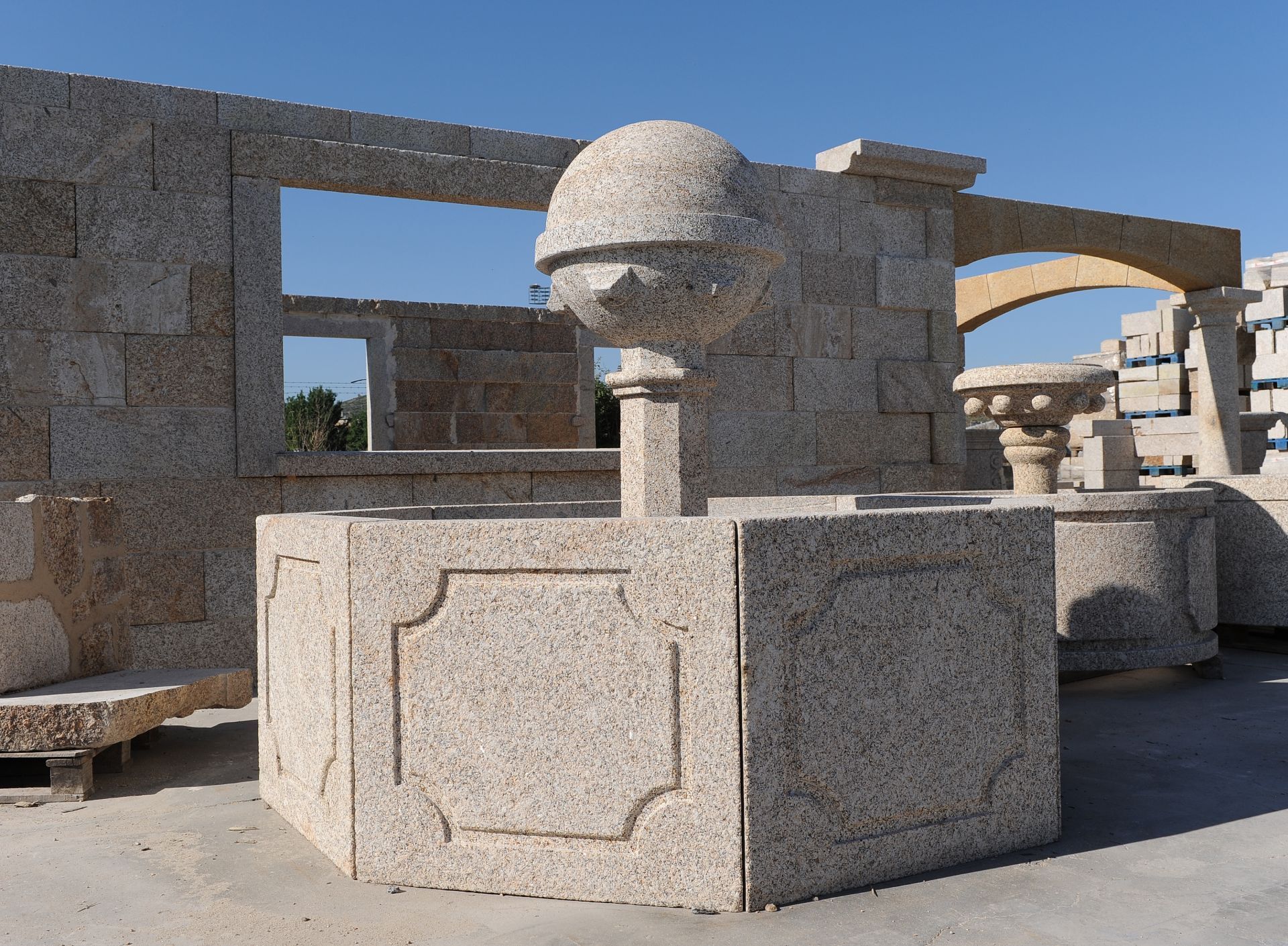 En un almacén de materiales de construcción al aire libre se encuentra una fuente de piedra de color claro con una esfera central.