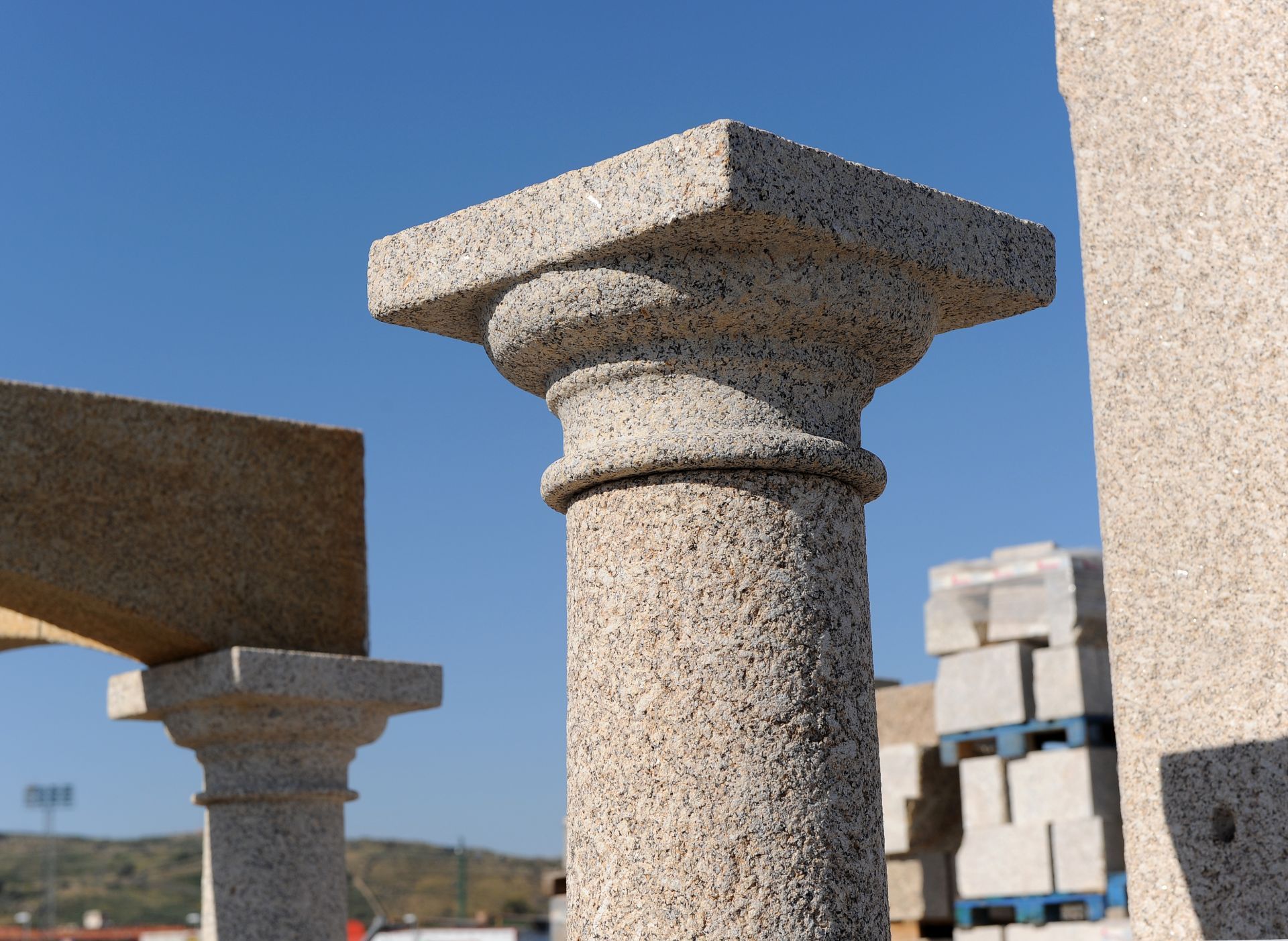 Columnas de piedra toscamente labradas con capiteles ornamentados se alzan contra un cielo azul despejado en una zona de construcción al aire libre.