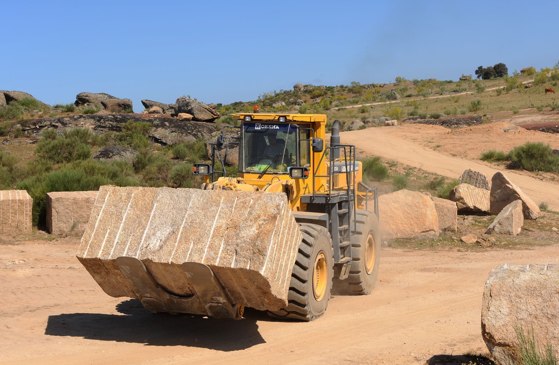 Una pala cargadora amarilla transporta un gran bloque de piedra cortada a través de una polvorienta cantera bajo un cielo azul despejado.