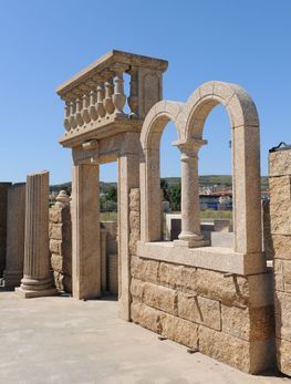 Estructuras arquitectónicas de piedra de color tostado, que incluyen columnas, ventanas arqueadas y una balaustrada, se alzan al aire libre contra un cielo azul.