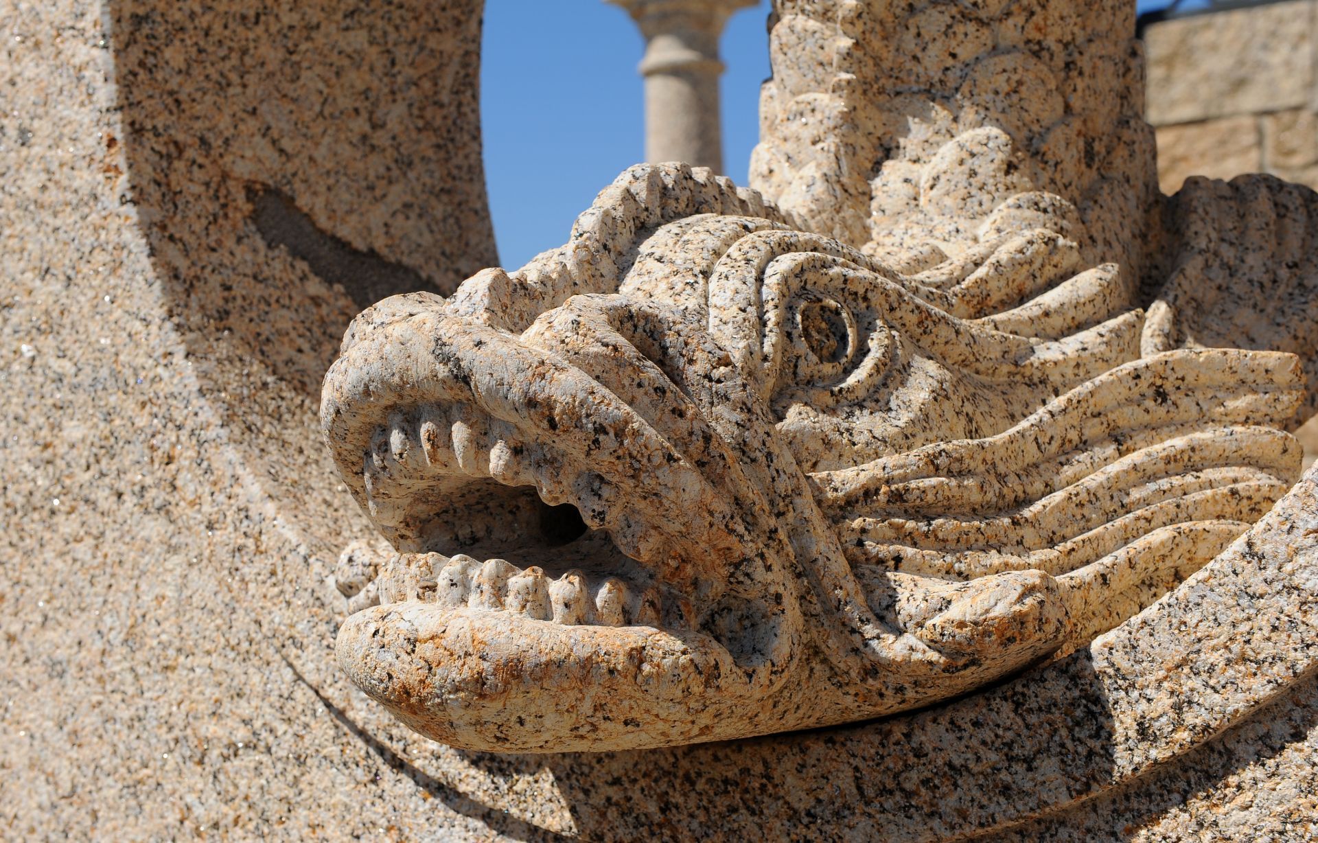 Primer plano de una escultura de una cabeza de dragón tallada en piedra de granito con la boca abierta y los dientes visibles, contra un cielo azul.