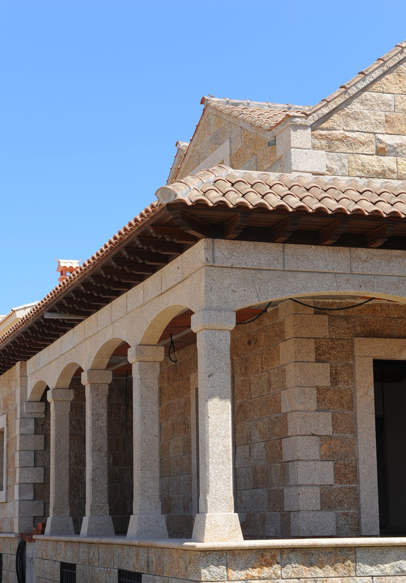 Un edificio de piedra con un pórtico abovedado cubierto y columnas bajo un tejado de tejas marrones, con un cielo azul despejado de fondo.