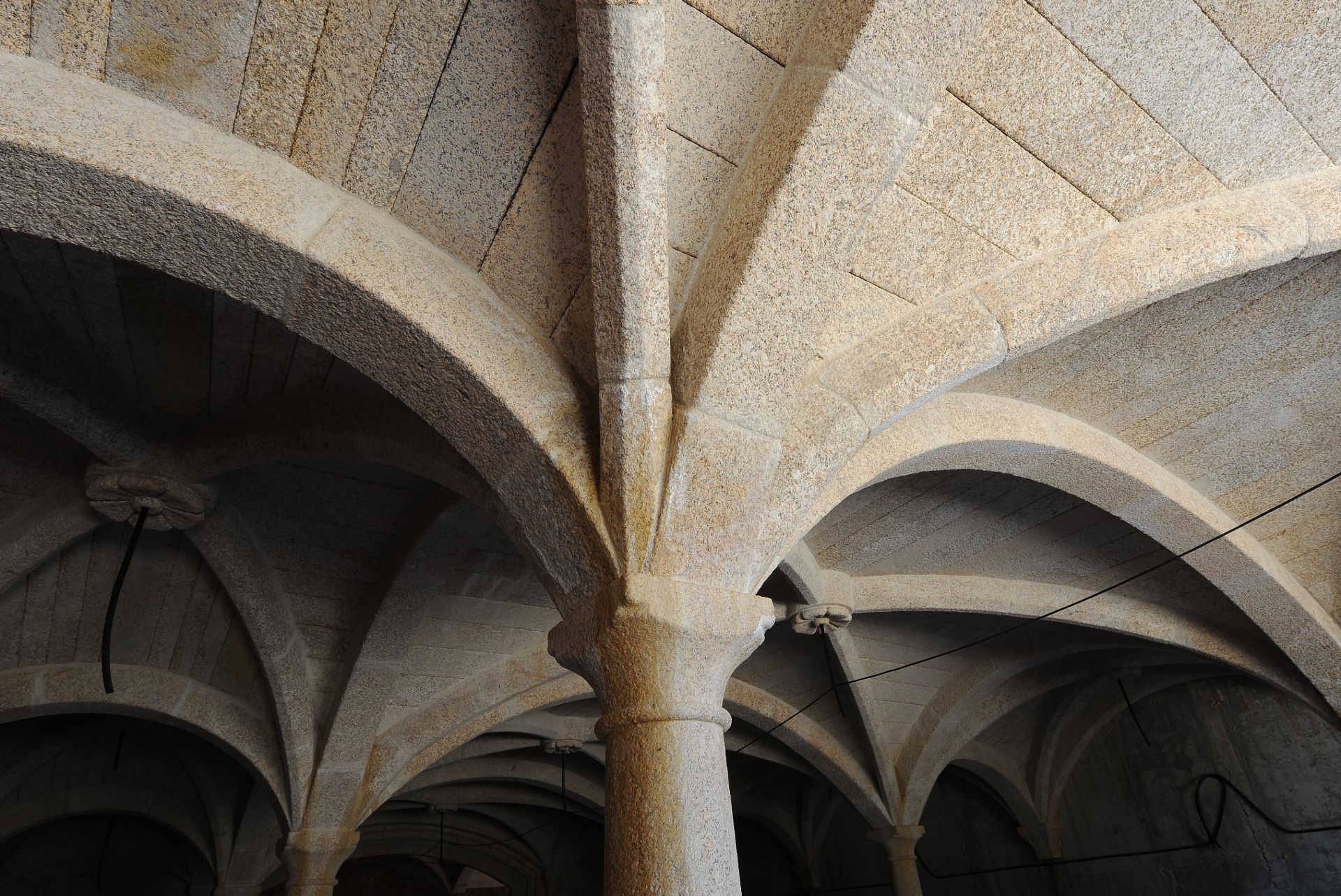Techo abovedado de piedra con nervaduras y arcos que se entrecruzan, sostenido por una columna central, en un interior histórico con iluminación tenue.
