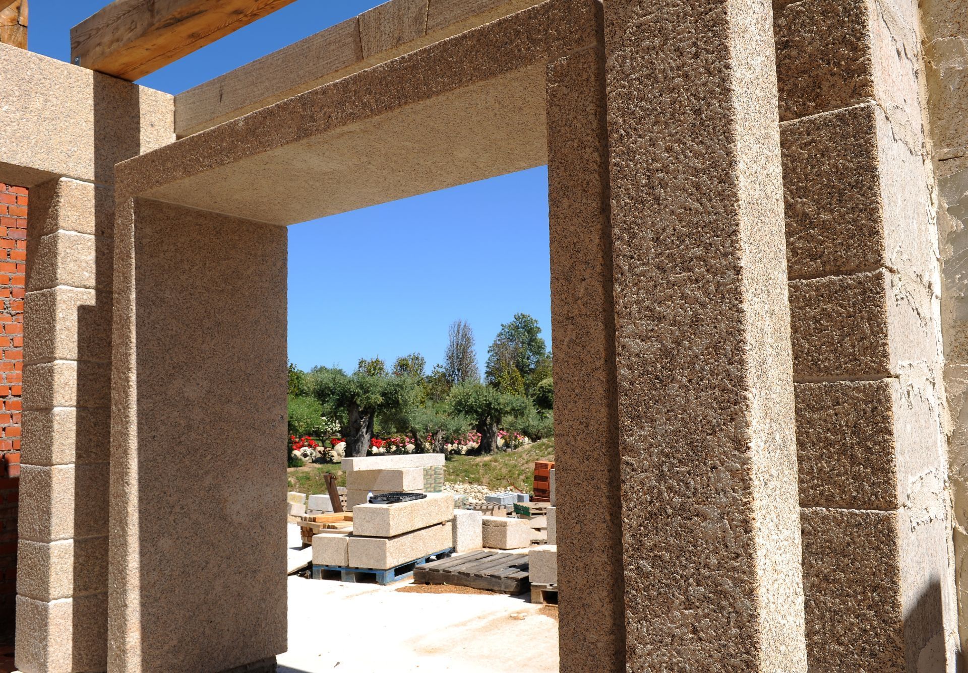 Una puerta en construcción, enmarcada por bloques de piedra texturizada, que da a la vista de un soleado patio exterior con ladrillos apilados.