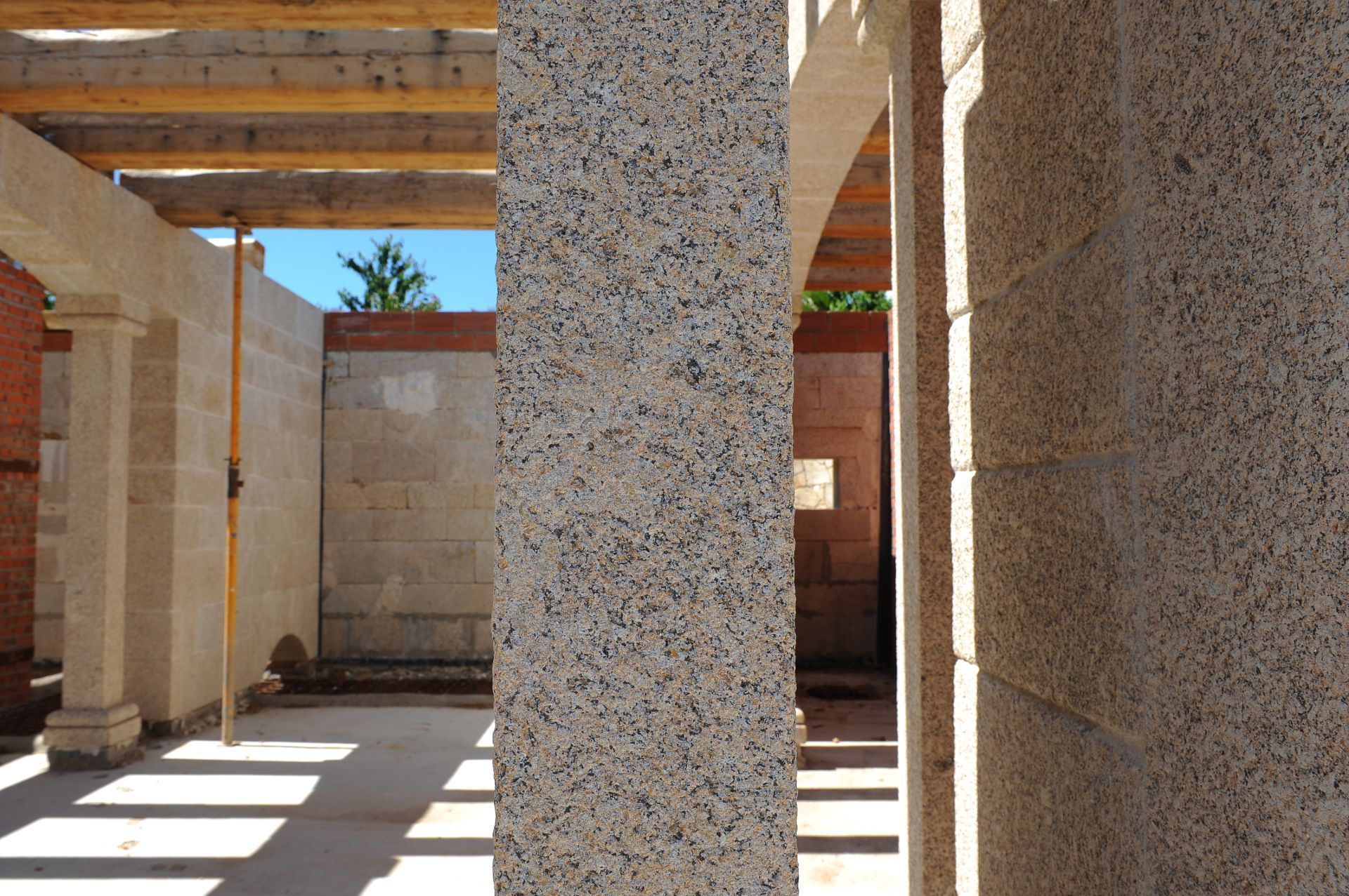 Edificio en construcción con columnas y paredes de granito y vigas de madera a la vista en el techo, bajo un cielo azul.