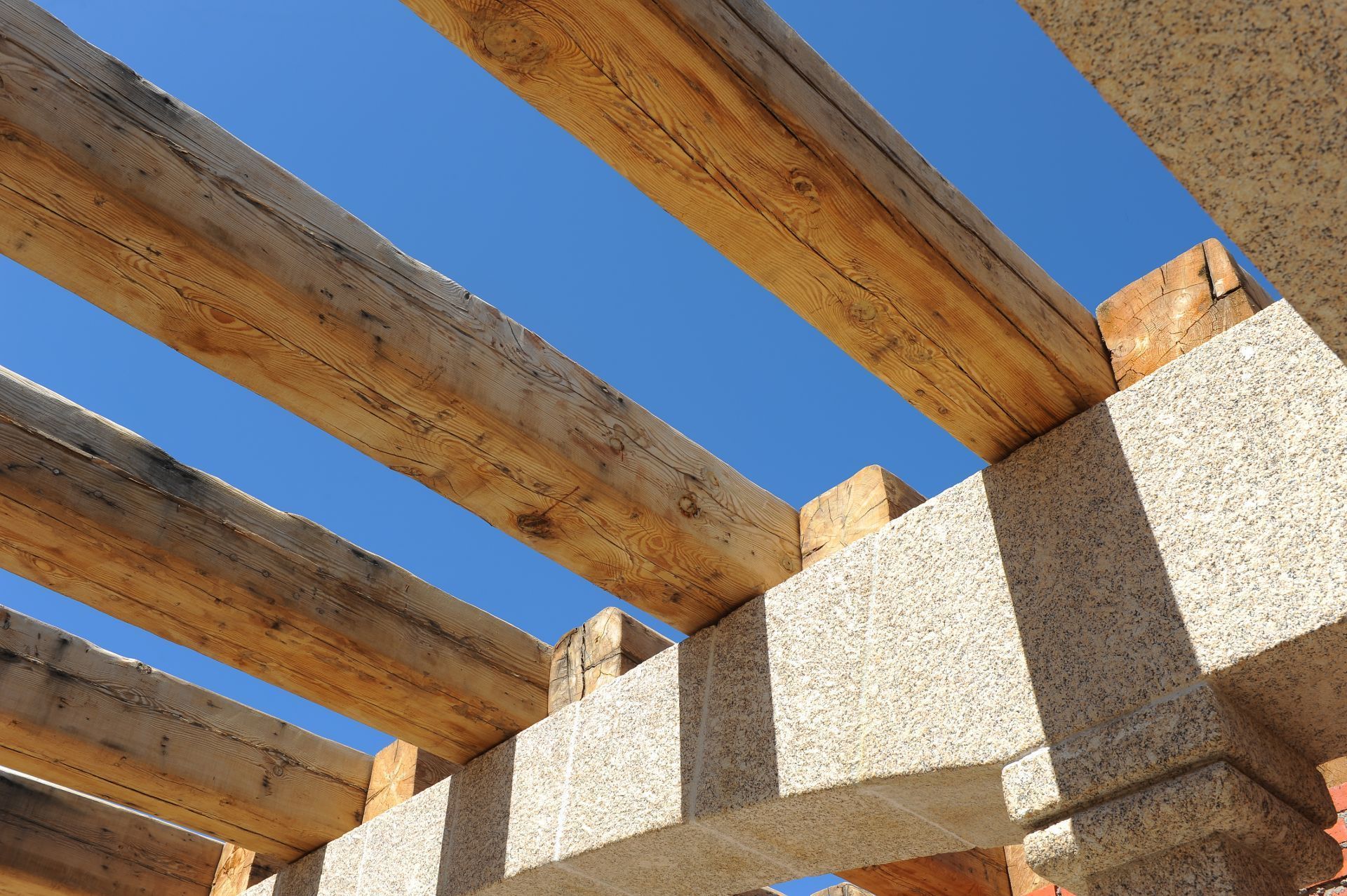 Vista desde un ángulo bajo de las vigas de madera de una pérgola que descansan sobre una estructura de hormigón texturizado, con un cielo azul despejado de fondo.