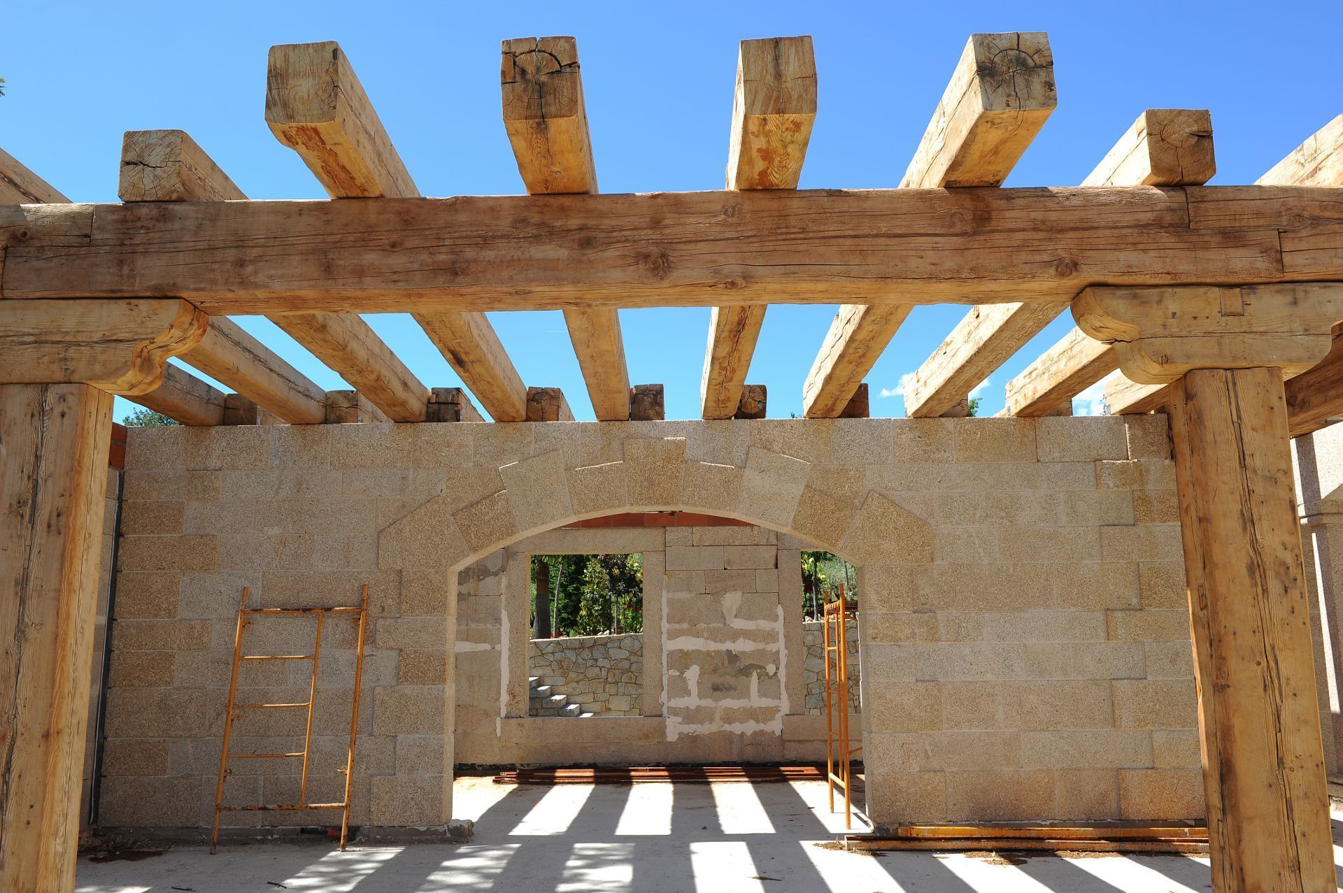 Un edificio de piedra parcialmente construido, con pesadas vigas de madera en el techo y una entrada central arqueada.