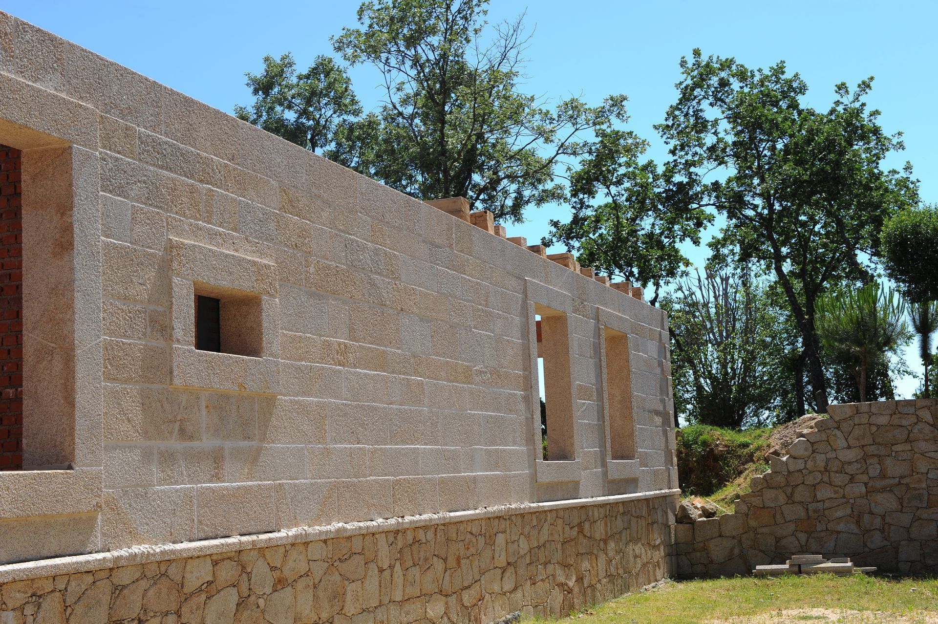 Un muro de piedra de color claro en construcción, con aberturas para ventanas, con un telón de fondo de árboles y un cielo azul.