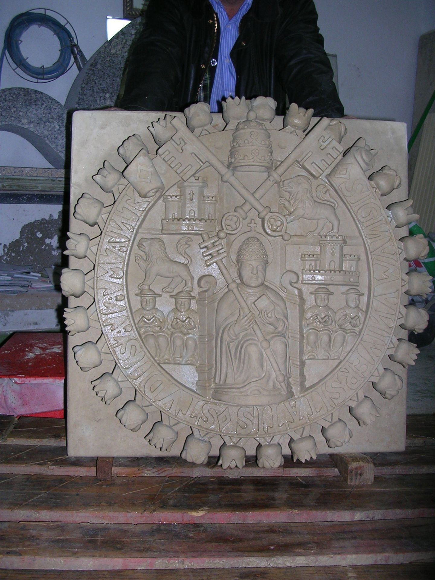 Una escultura en piedra que representa un escudo de armas papal con una figura central, llaves y una tiara, sostenida por una persona en un taller.
