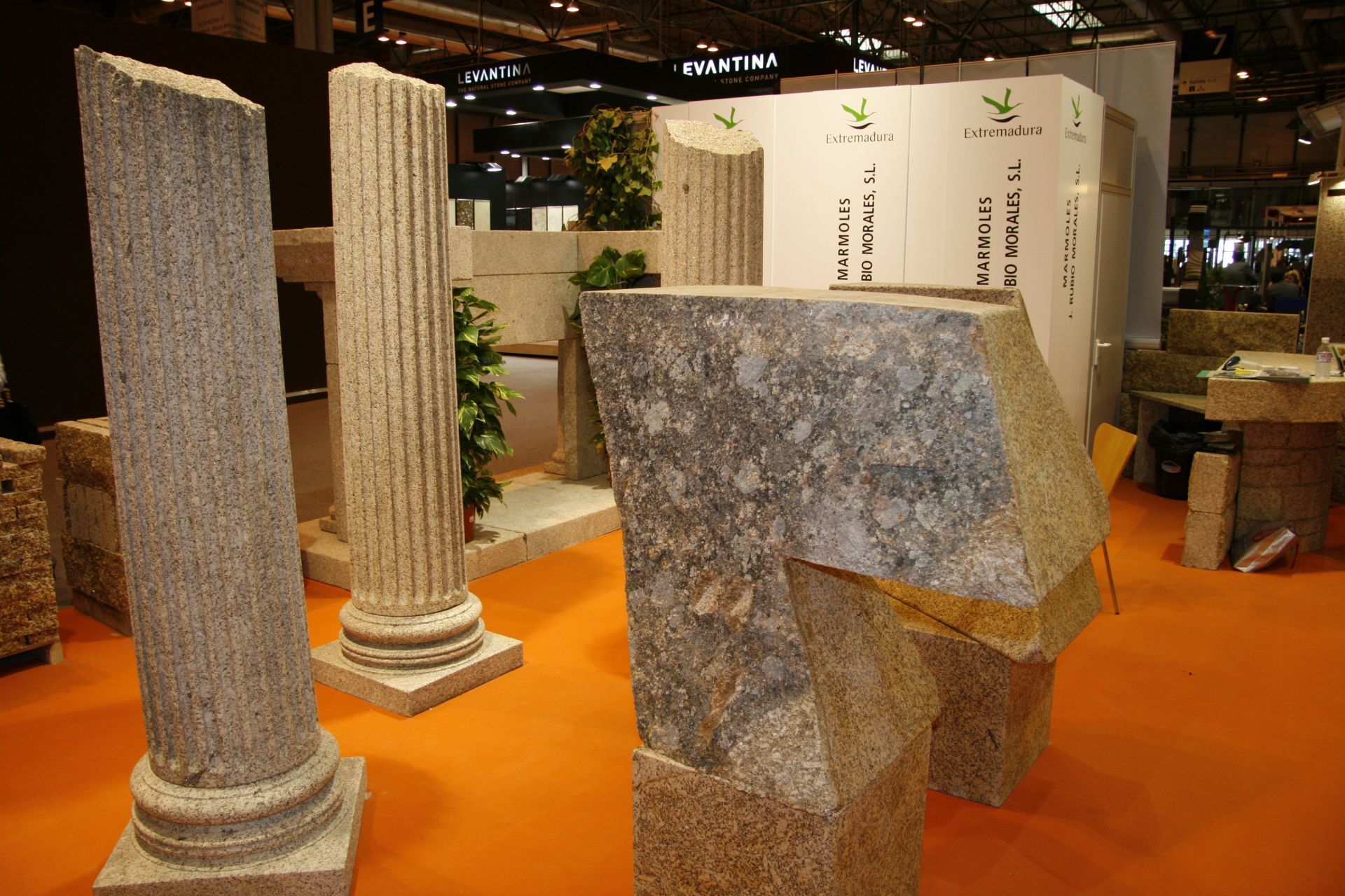 Exhibiciones de columnas de piedra tallada y esculturas abstractas sobre un suelo naranja en un entorno de feria comercial.