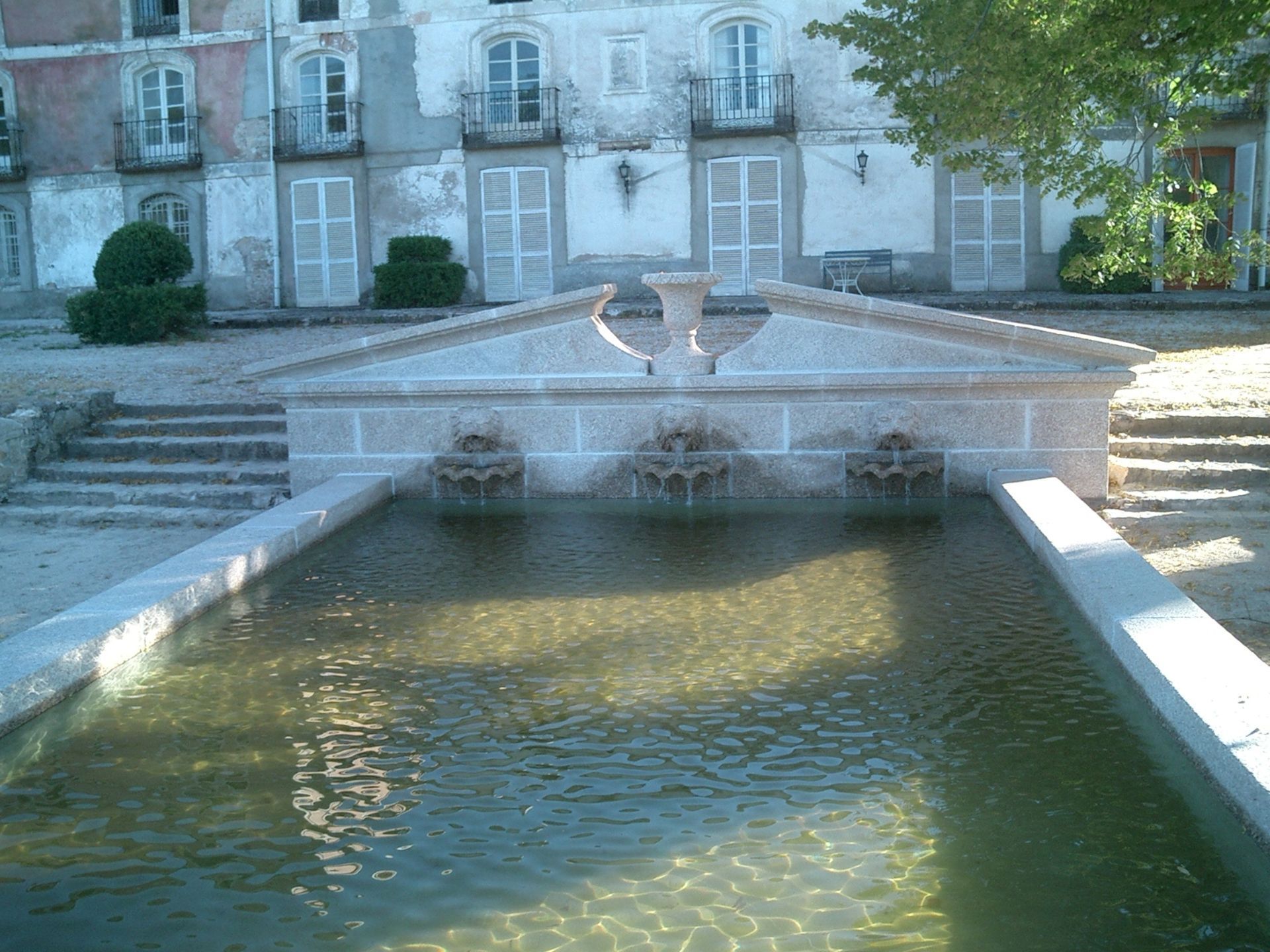 Una fuente de piedra con tres caños tallados acumula agua frente a un edificio antiguo de varias plantas con contraventanas blancas.