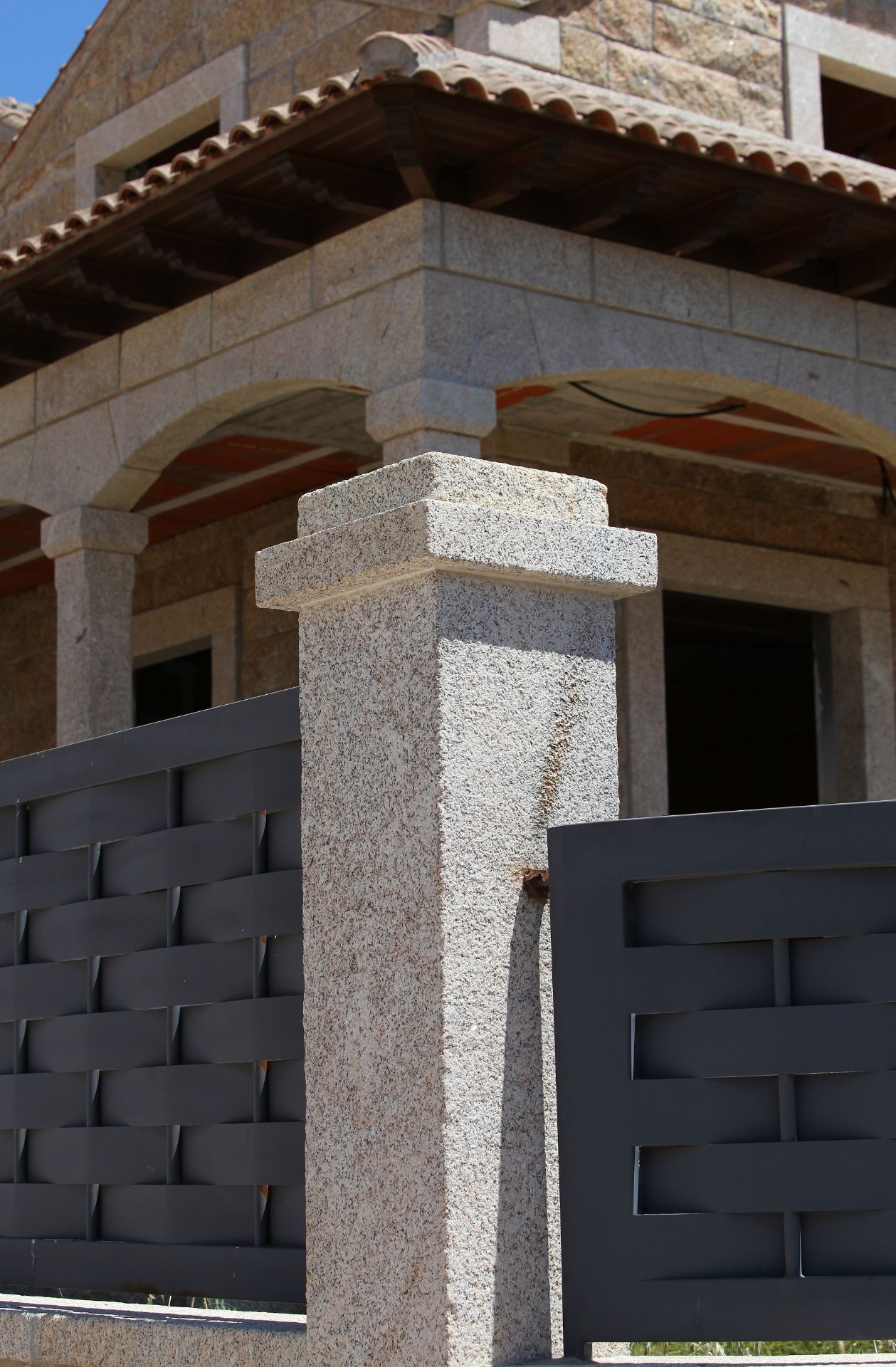 Un pilar de piedra texturizada se alza frente a una casa con techo de tejas y valla metálica con diseño de cesta.