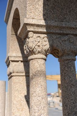 Primer plano del capitel de una columna de piedra tallada con motivos de hojas, que sostiene una estructura arqueada contra un cielo azul.