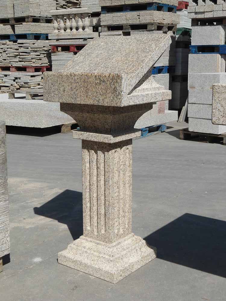 Un atril de piedra de granito color tostado con una base de columna estriada, expuesto al aire libre en un almacén de materiales de construcción.