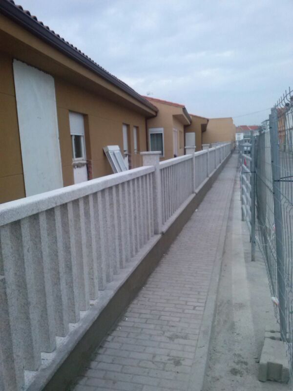 Un sendero pavimentado discurre entre un edificio residencial marrón con una balaustrada de piedra blanca y una valla de malla metálica.