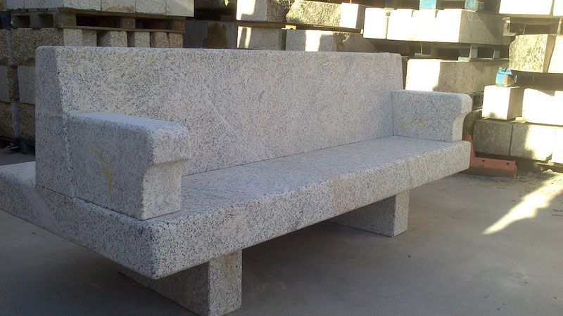 Un banco de piedra de parque con respaldo alto y reposabrazos, situado en una zona de almacén llena de bloques de piedra.